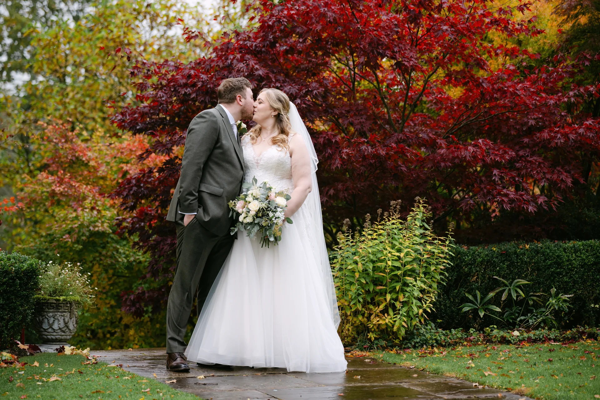A newlywed couple shares a tender kiss on a paved path surrounded by vibrant autumn foliage and striking red maple trees.