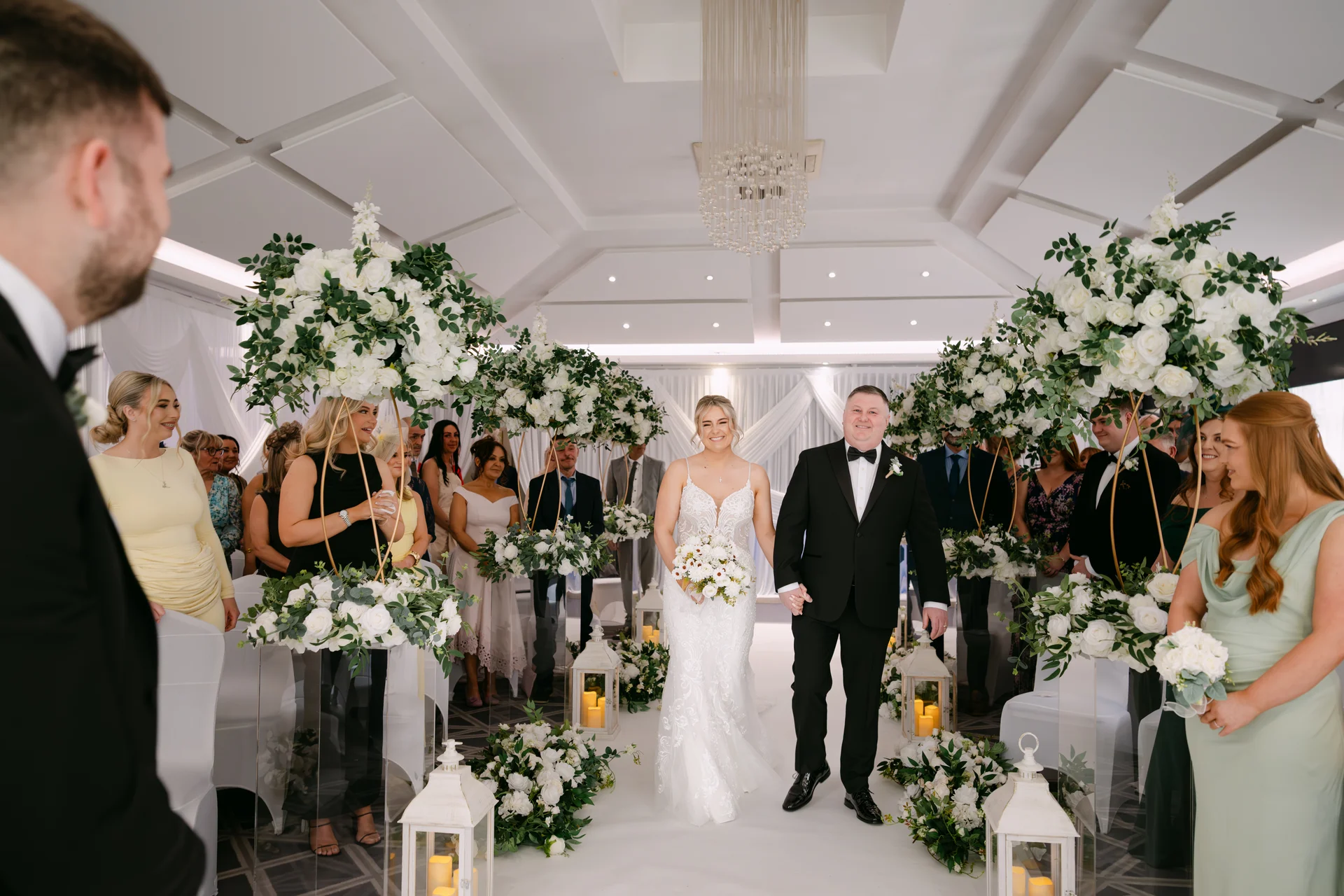 A smiling bride and groom walk hand in hand down the aisle after their civil ceremony, flanked by large white floral arrangements and seated guests.