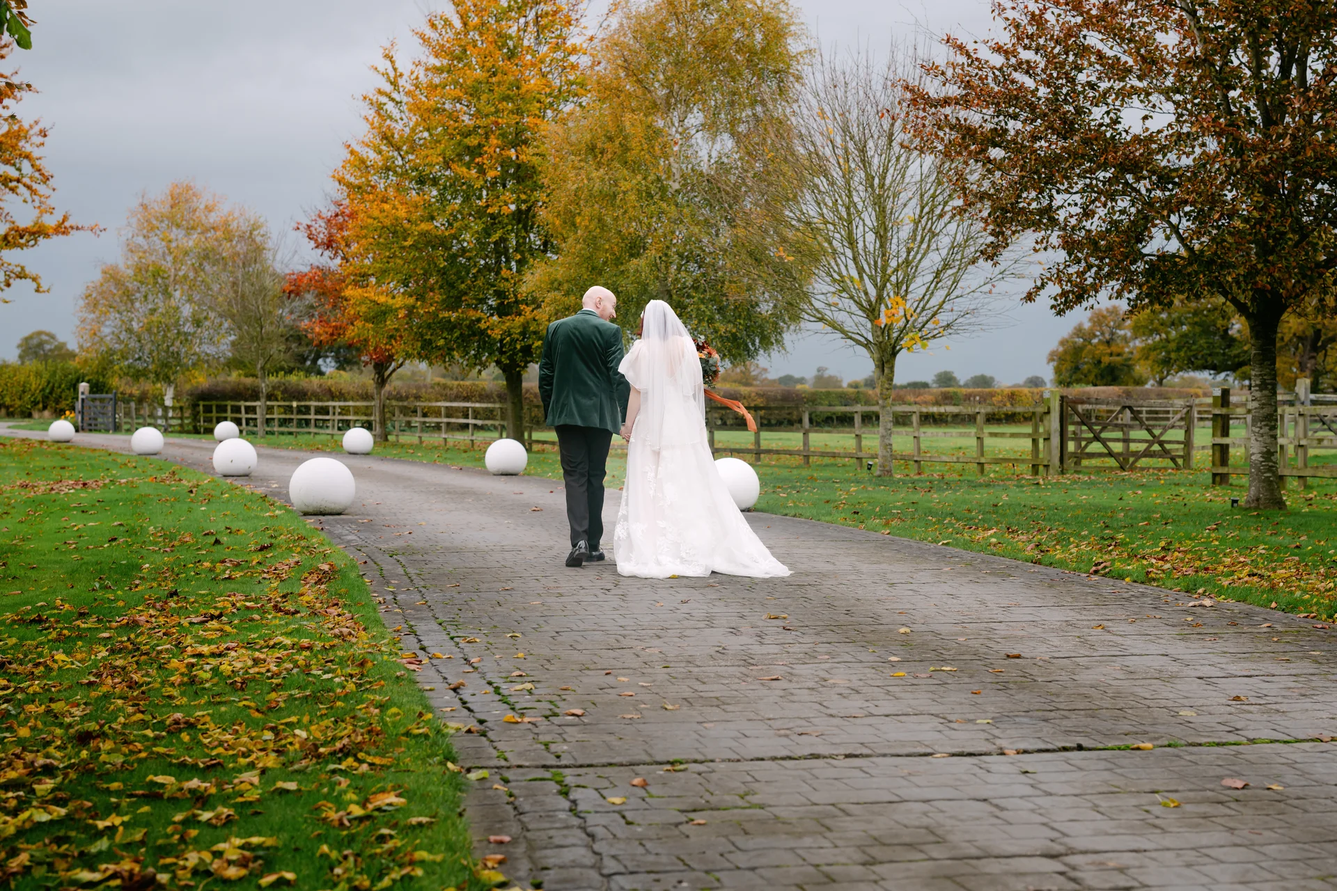 A newlywed couple walks hand in hand away from the camera down a stone driveway lined with autumn trees and glowing white orb lights.