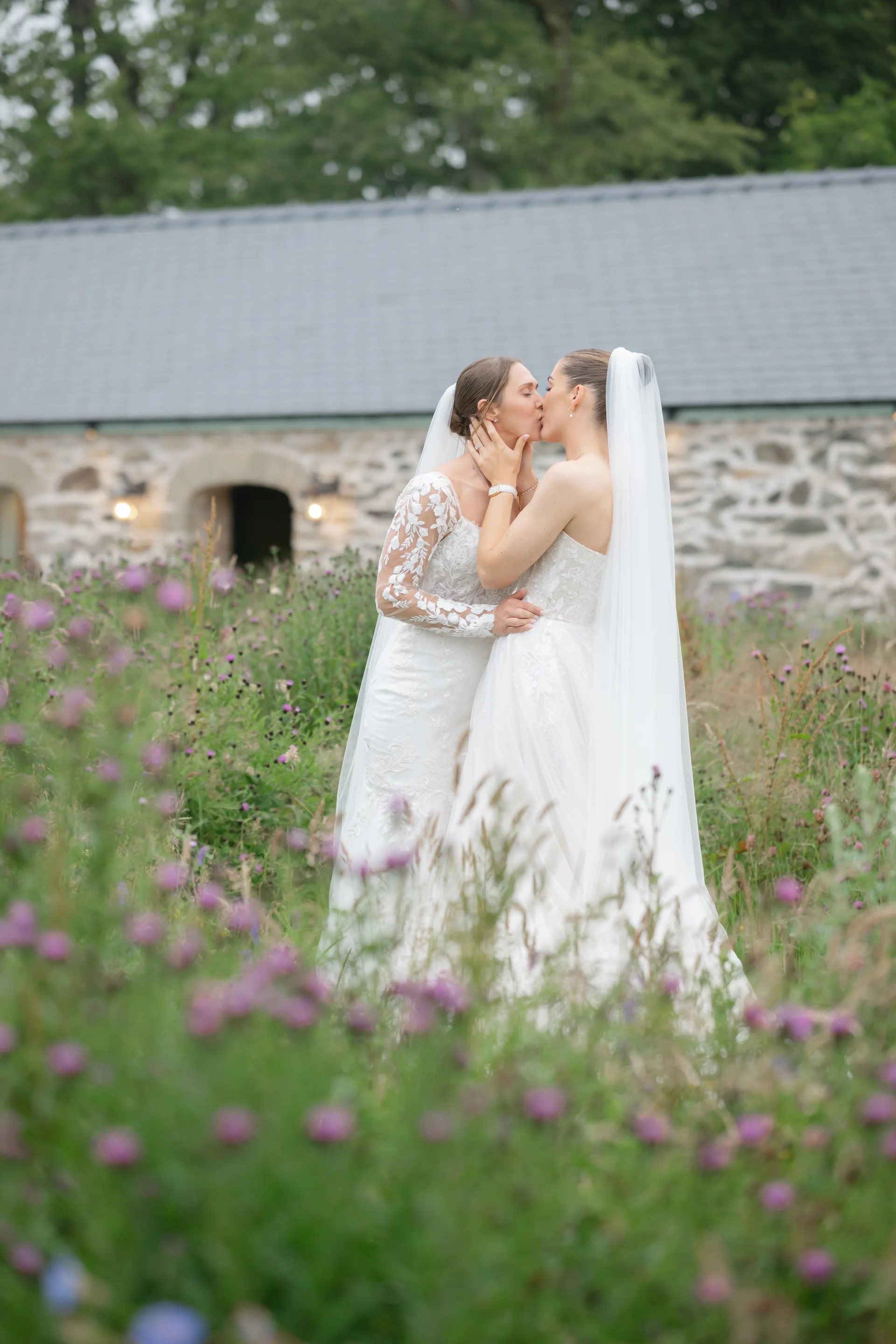 Two brides share a tender kiss amidst a vibrant wildflower meadow with a rustic stone barn in the background.