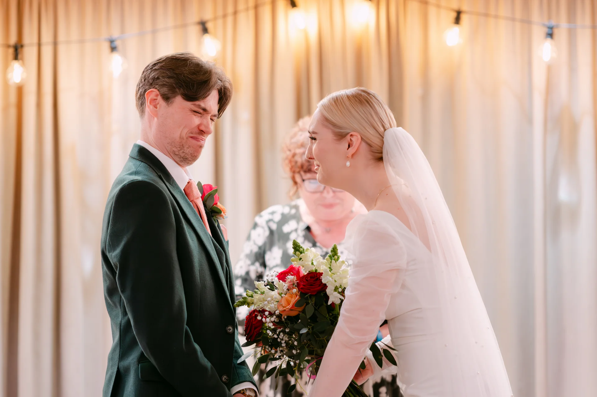 An emotional groom tears up while standing opposite his laughing bride, who holds a vibrant hand-tied bouquet during their wedding ceremony.