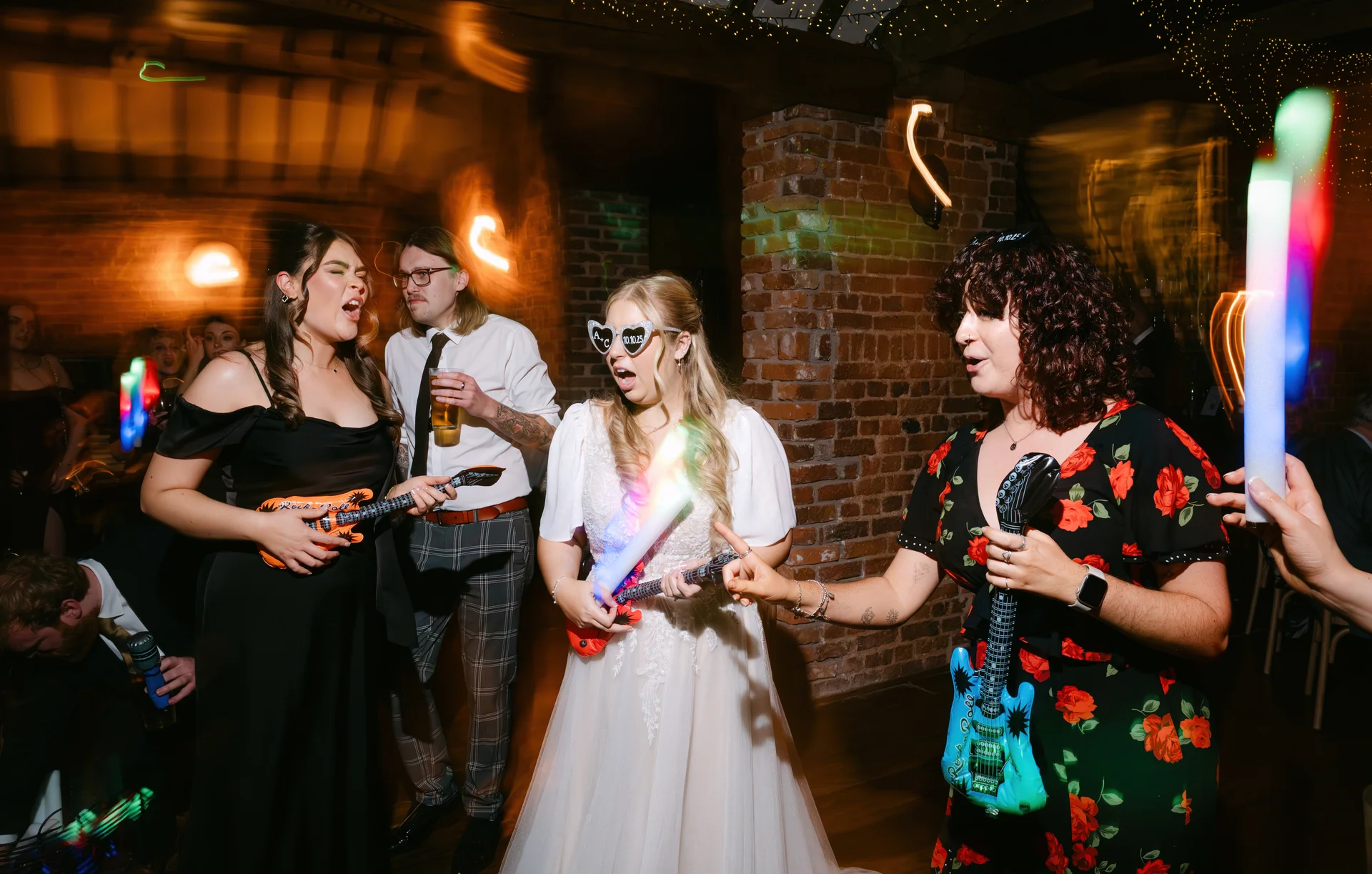 A lively party scene on a dance floor with guests dancing enthusiastically, holding inflatable guitars and glowing sticks in front of an exposed brick wall.