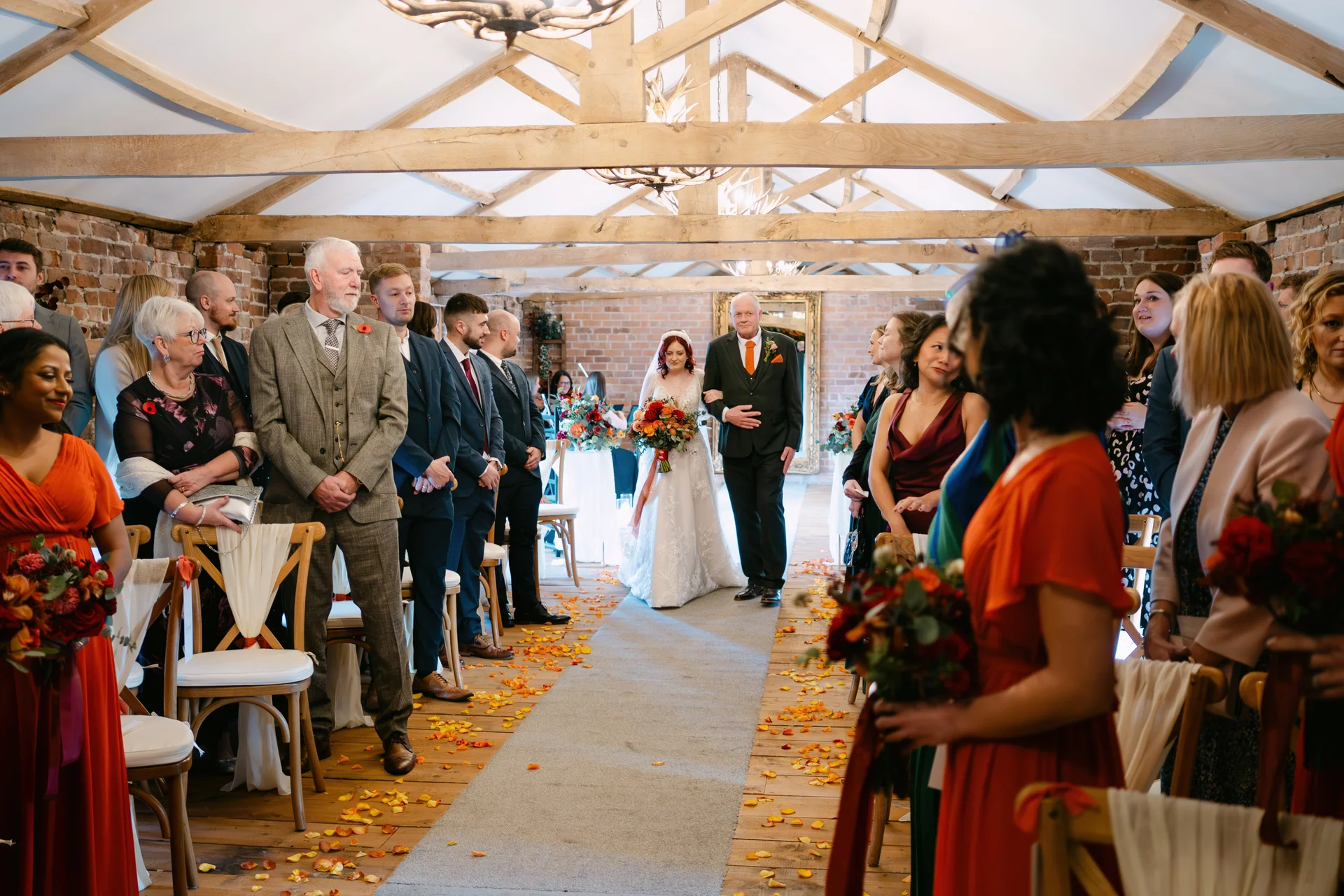 The bride is walking down the aisle with her father in a rustic barn ceremony venue lined with seated guests and autumnal petal decor.