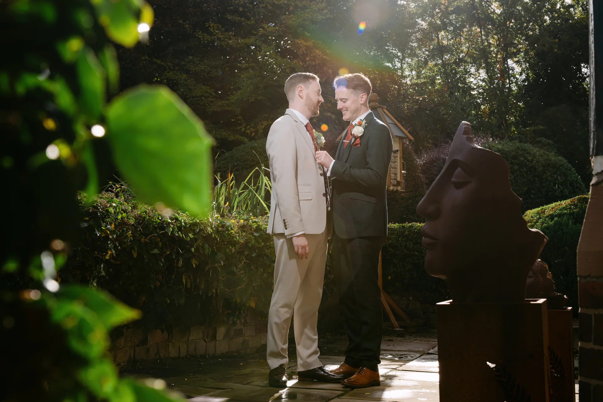 Two grooms stand together in a sunny garden, framed by foliage in the foreground with a dramatic lens flare from behind.