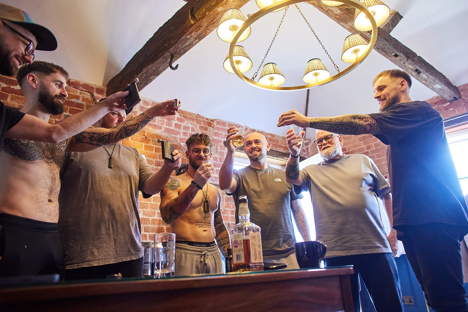 A group of groomsmen raise a celebratory toast with drinks and hip flasks in a rustic room with exposed brick and wooden beams during wedding morning preparations.