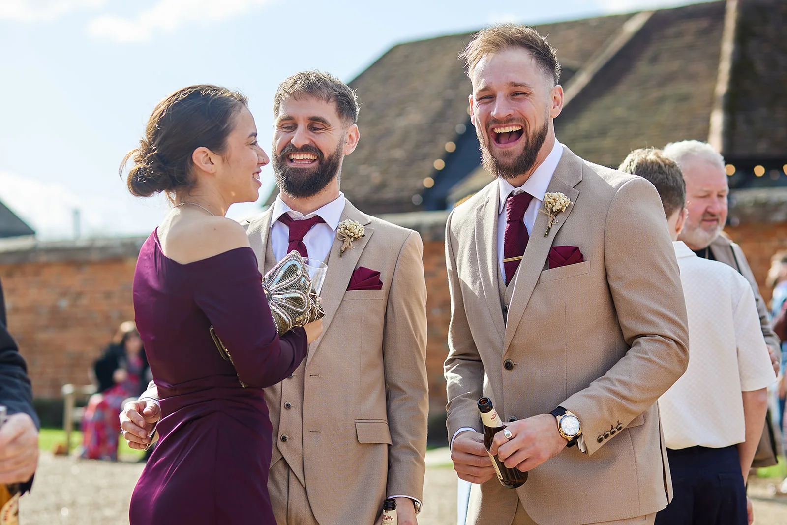 A group of wedding guests share a cheerful moment outdoors in bright sunlight, wearing beige suits with burgundy ties and a plum dress.