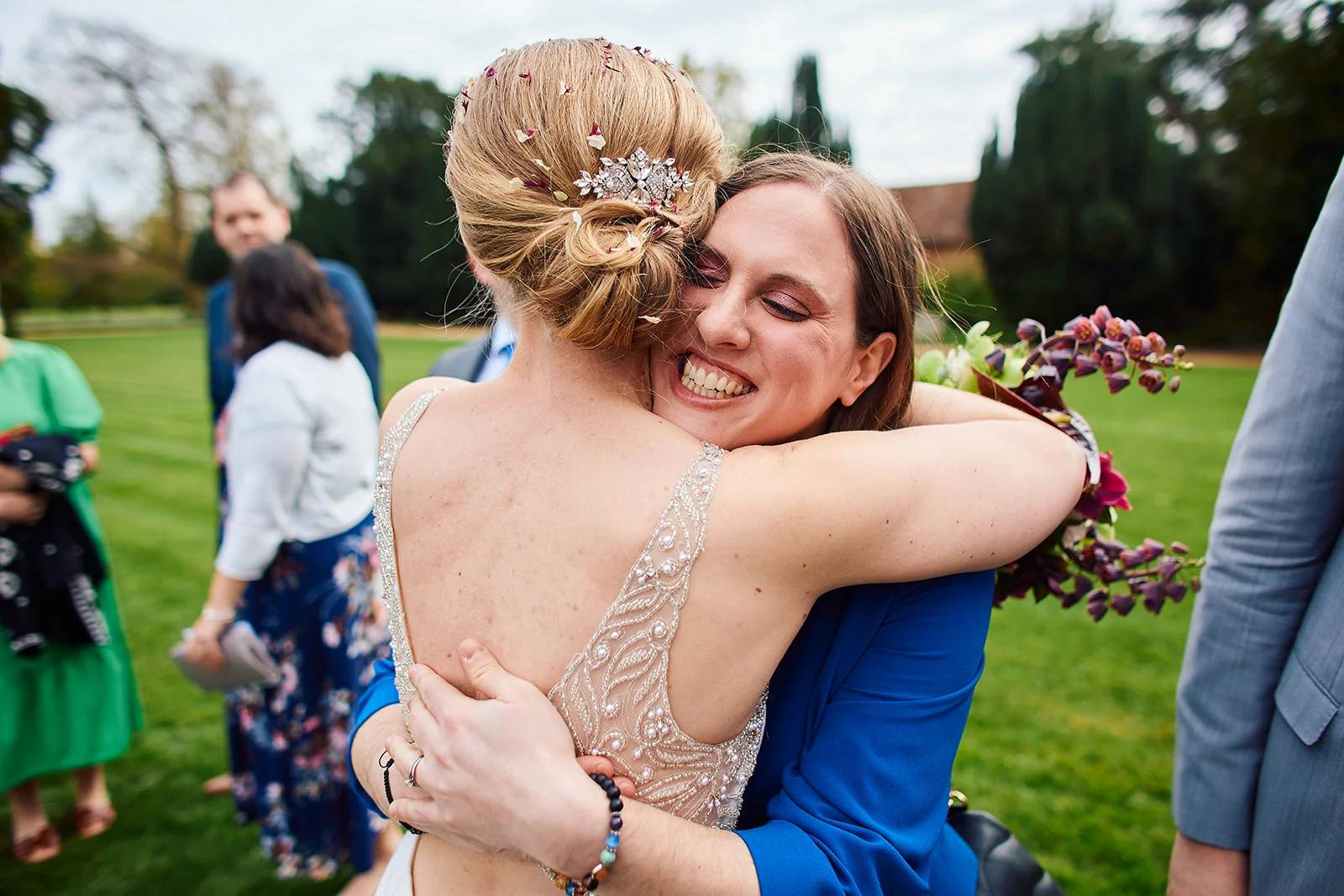 Two brides embrace joyfully outdoors in a garden setting, with confetti visible in one bride's blonde hair and wedding guests mingling in the background.