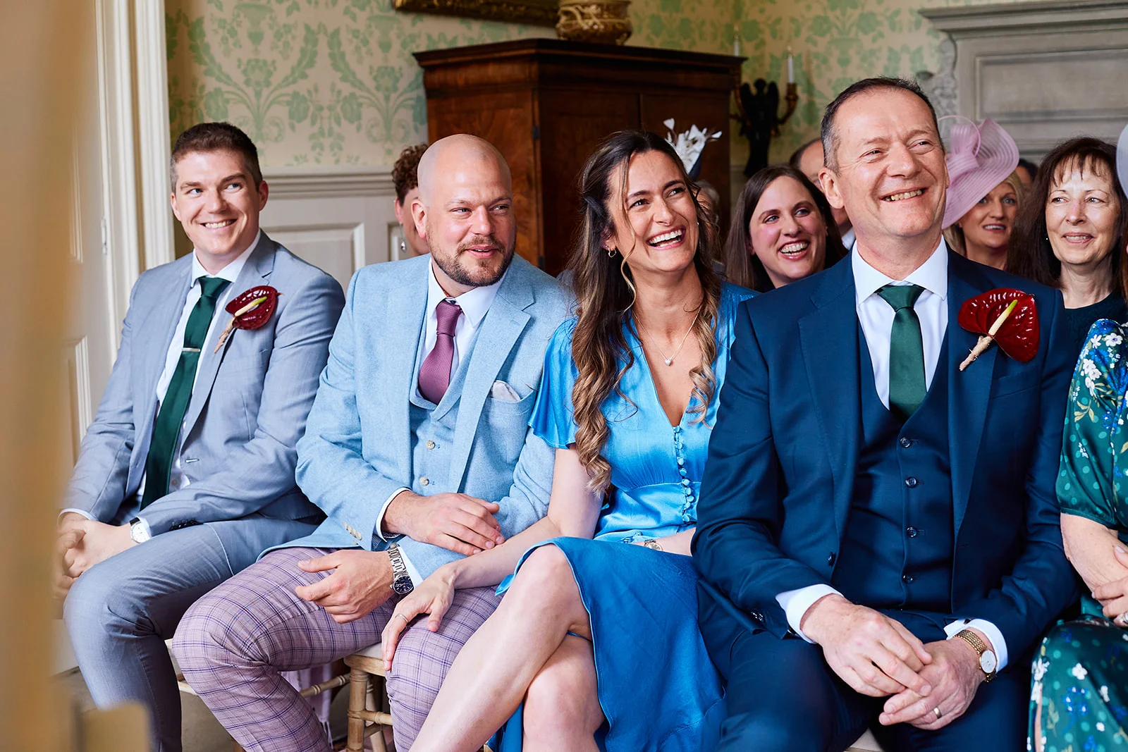 A group of wedding guests seated indoors, laughing happily during a ceremony inside a wood-panelled room.