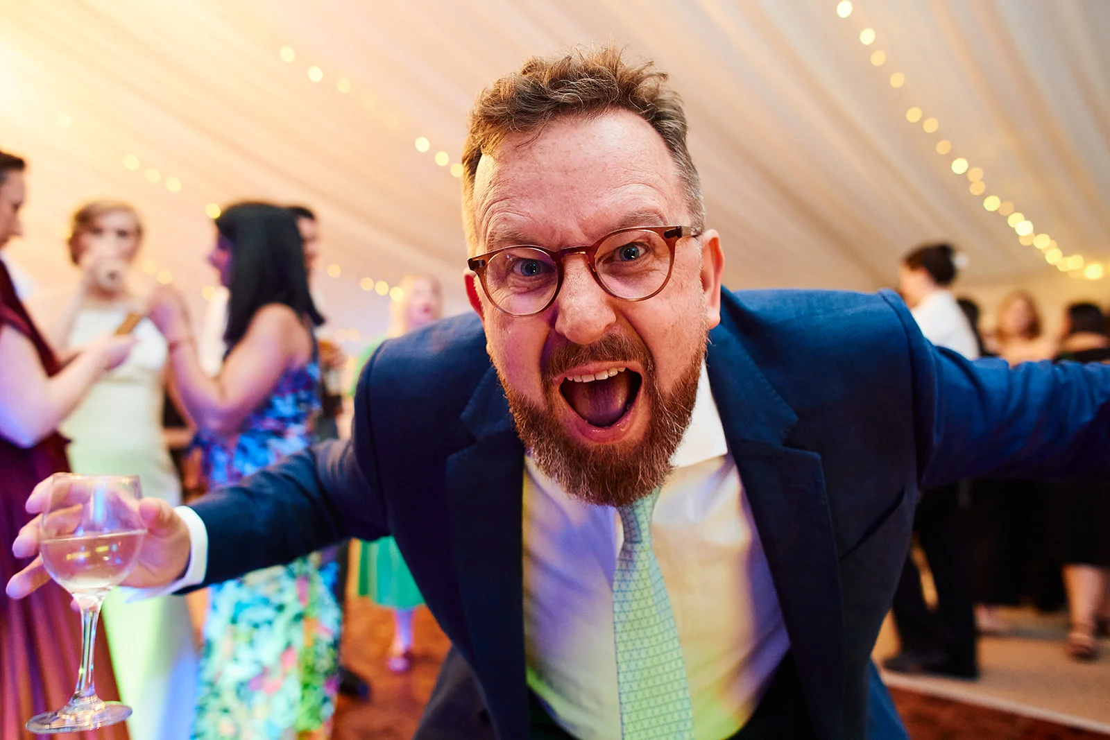 A joyous wedding guest in a blue suit leans playfully towards the camera holding a glass of wine inside a marquee lit by fairy lights.