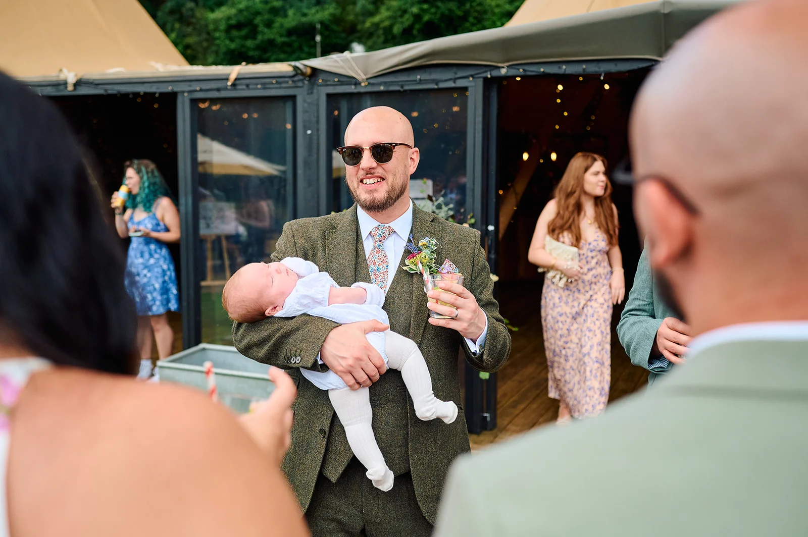 A smiling guest wearing sunglasses and a tweed suit holds a sleeping baby outside a wedding marquee during a summer celebration.