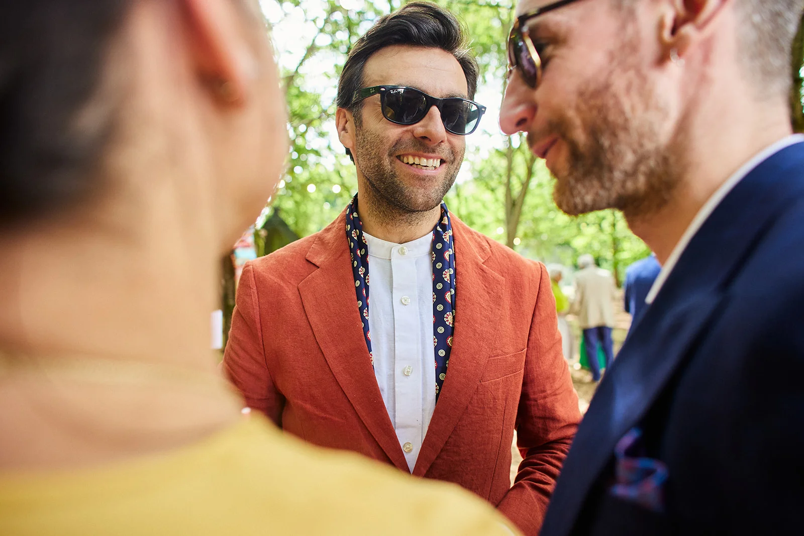 A smiling man wearing sunglasses and a terracotta suit jacket with a patterned neckerchief chats with guests outdoors on a sunny day.