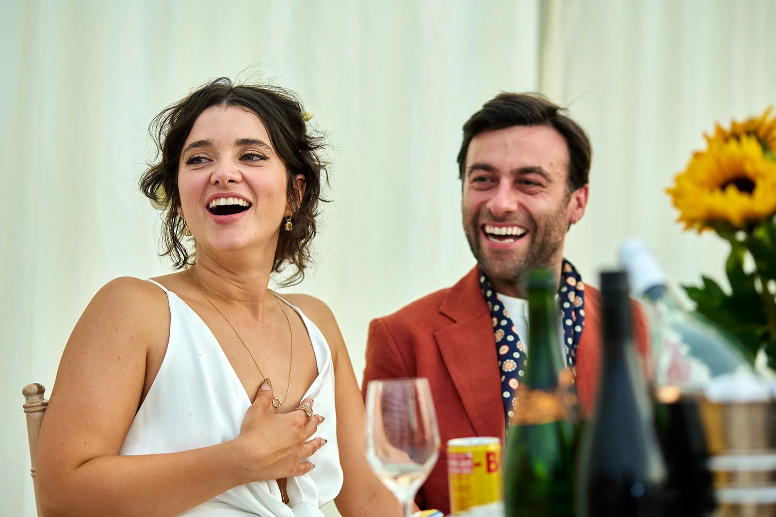 A newlywed couple laugh joyfully during wedding speeches, seated at a top table decorated with sunflowers and wine bottles.