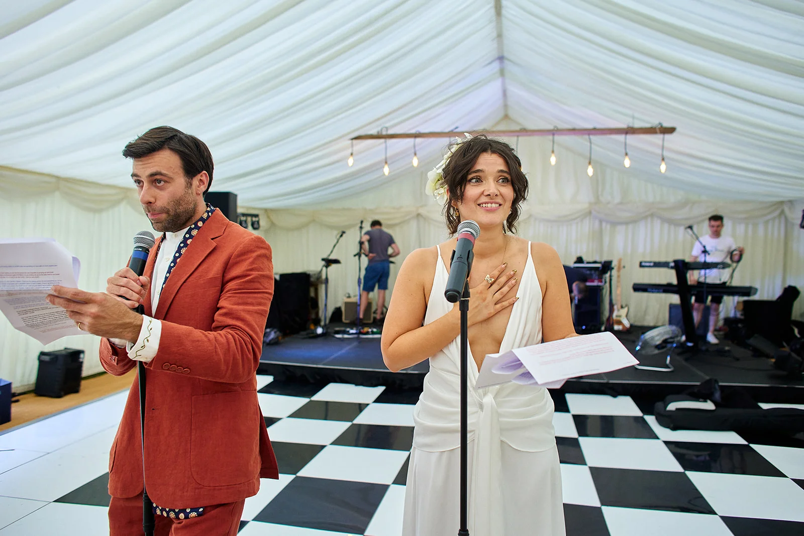 A bride and groom deliver their speeches holding microphones inside a marquee with a black and white checkerboard dance floor.