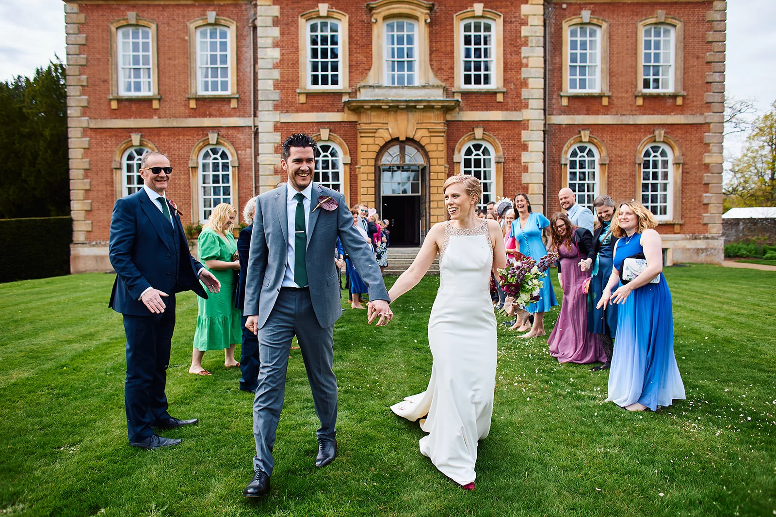 A joyful newlywed couple walk hand in hand across a manicured lawn in front of a grand brick manor house while guests cheer and celebrate.