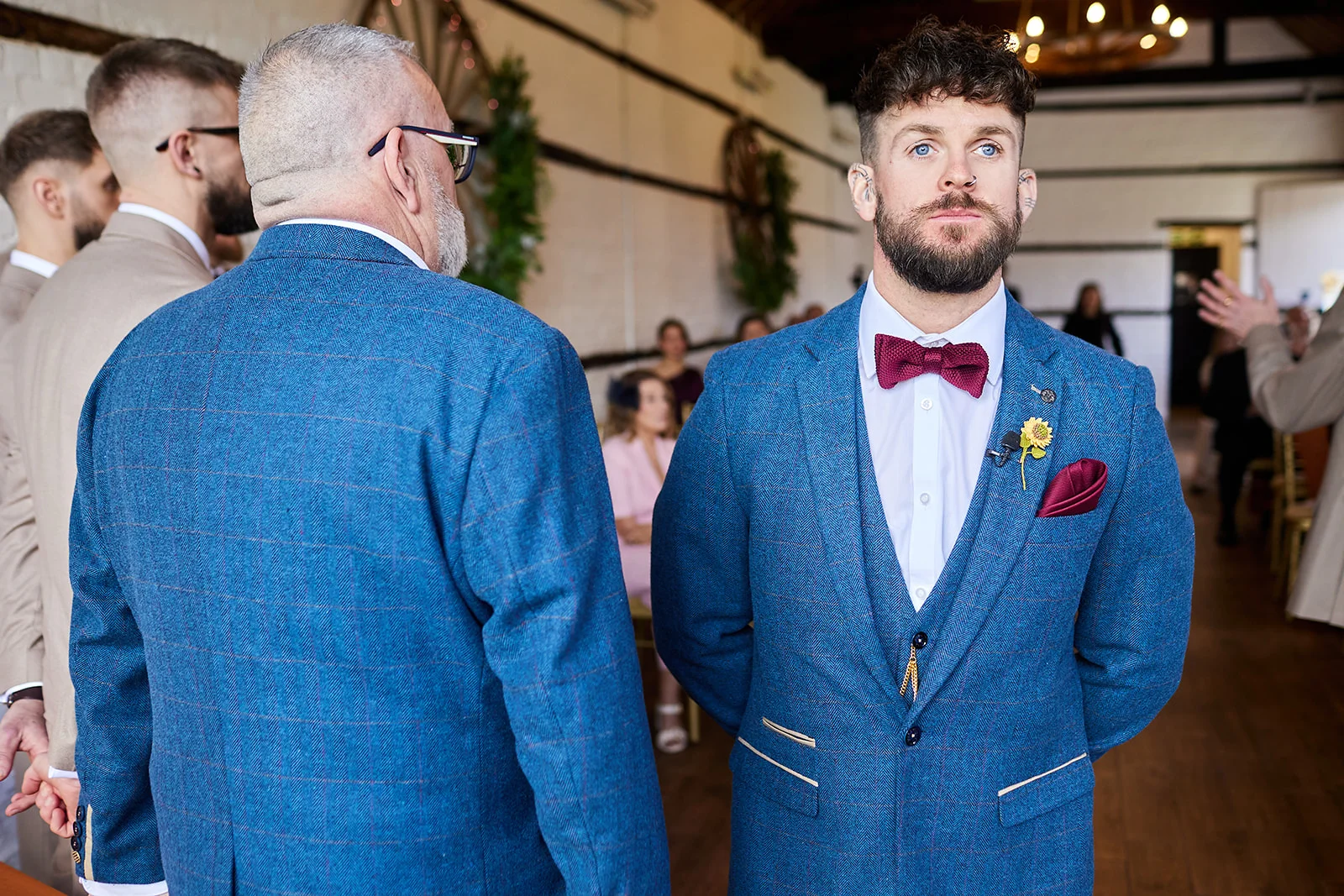 An emotional groom in a blue tweed suit with a burgundy bow tie waits at the altar inside a rustic barn venue.