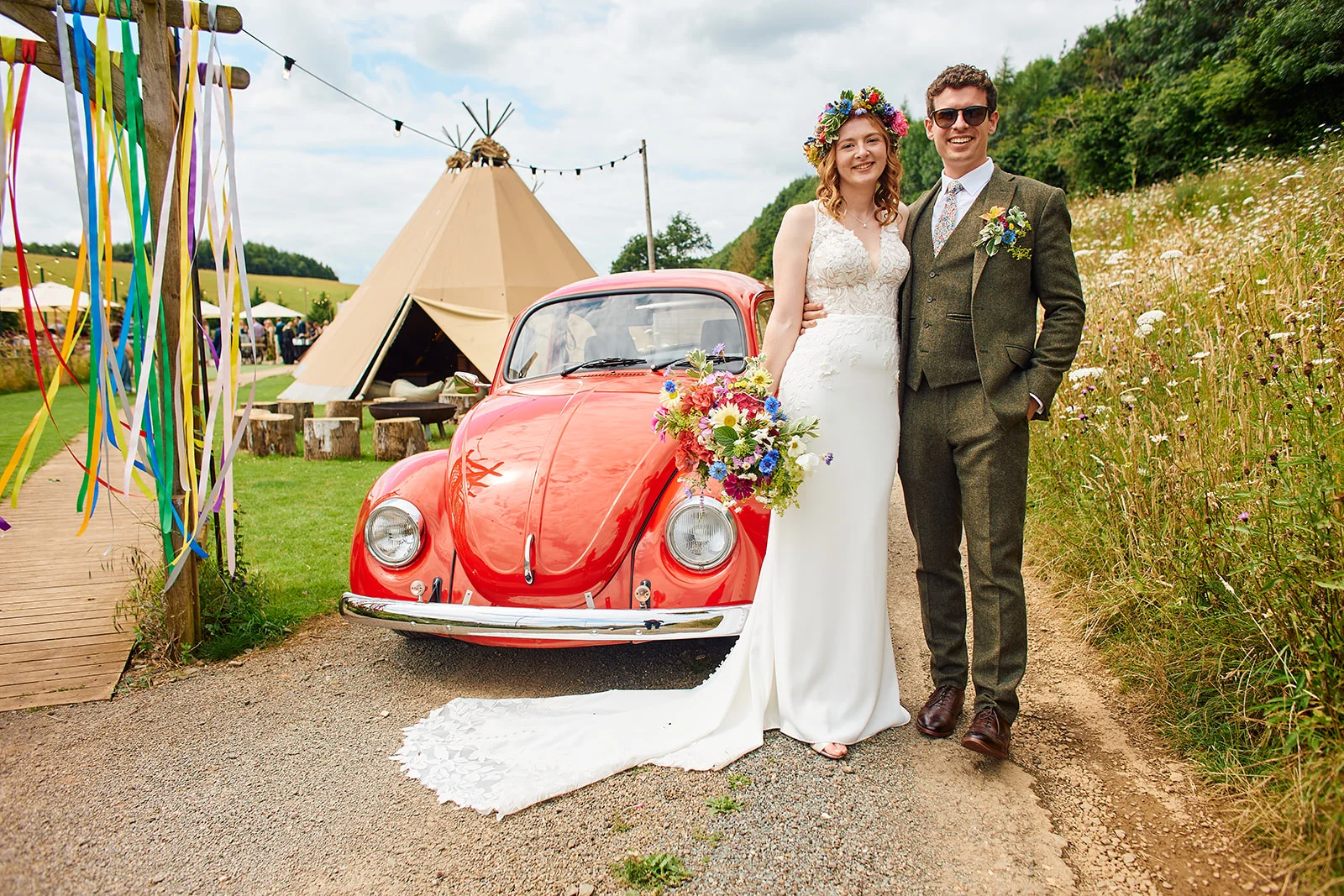 A smiling bride and groom pose next to a vintage red Volkswagen Beetle with a tipi and colourful ribbons in the background at an outdoor festival-style wedding.