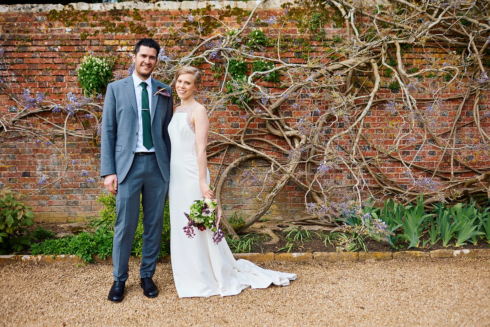 A smiling bride and groom stand together outdoors in front of a rustic brick wall covered with twisting wisteria branches, holding a trailing bouquet.