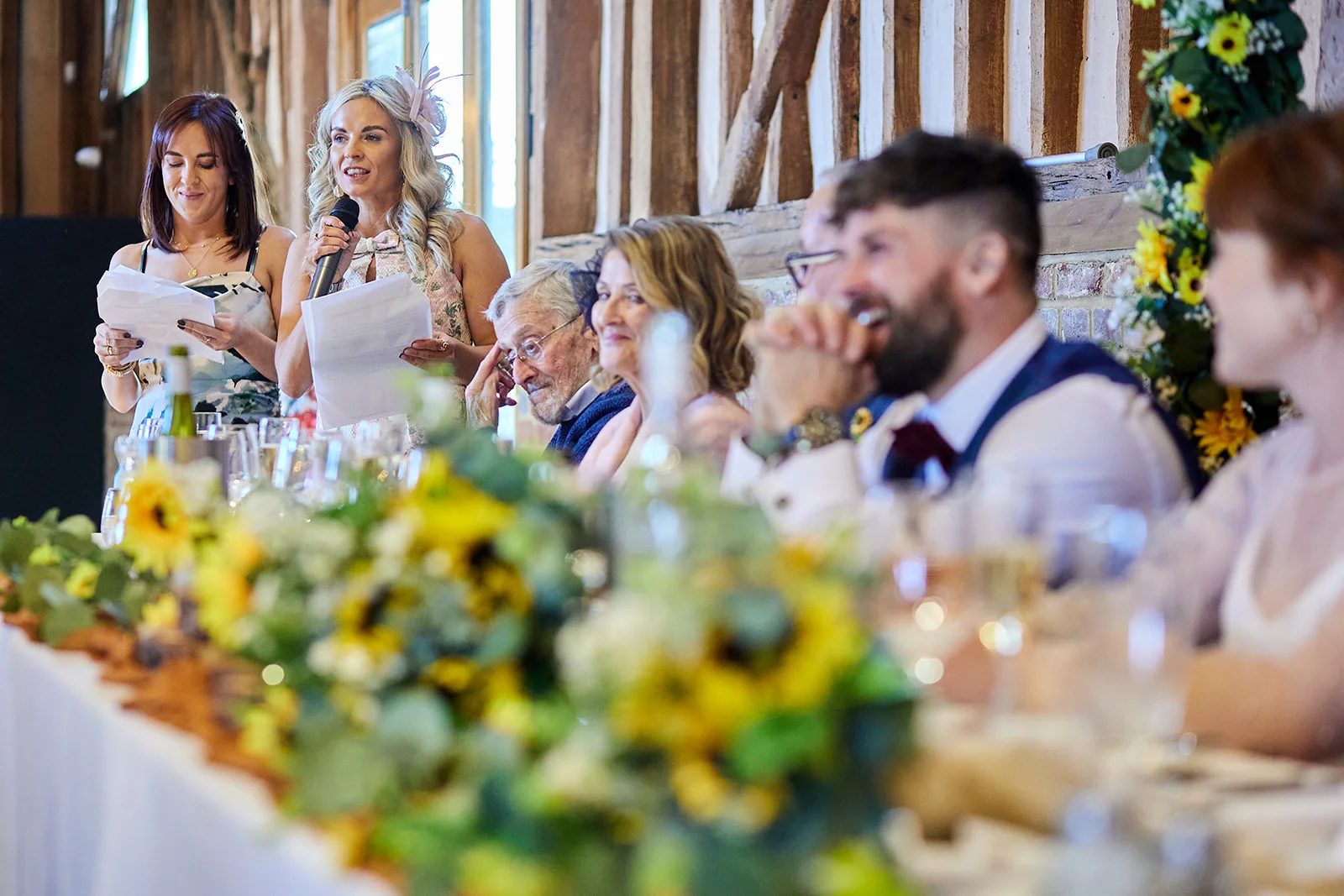 A smiling bride delivers a speech holding a microphone at the top table inside a rustic oak-framed barn wedding reception, flanked by wedding party members and guests enjoying the moment.