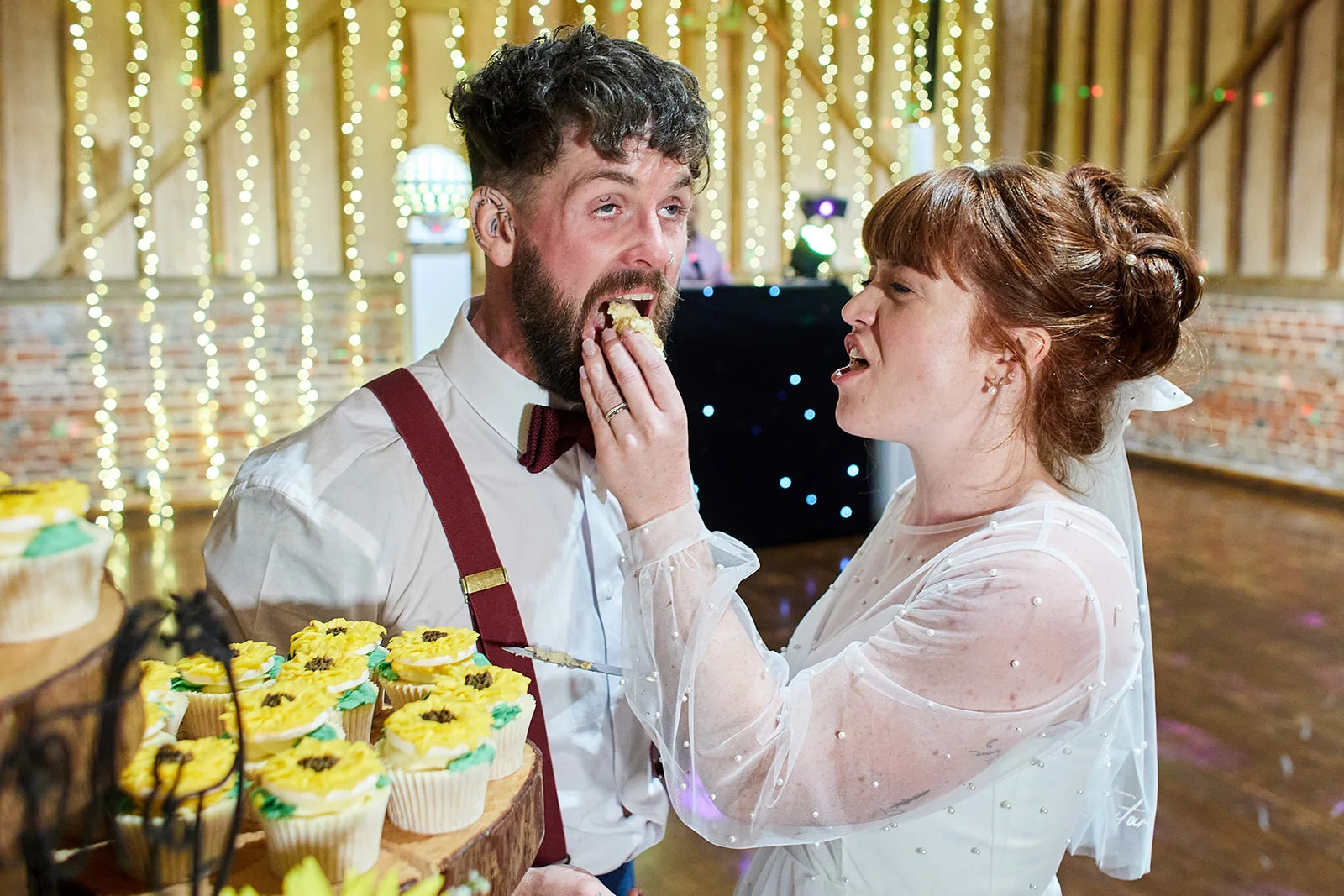 A newlywed couple playfully feed each other cupcakes inside a rustic barn decorated with fairy lights and exposed brick.