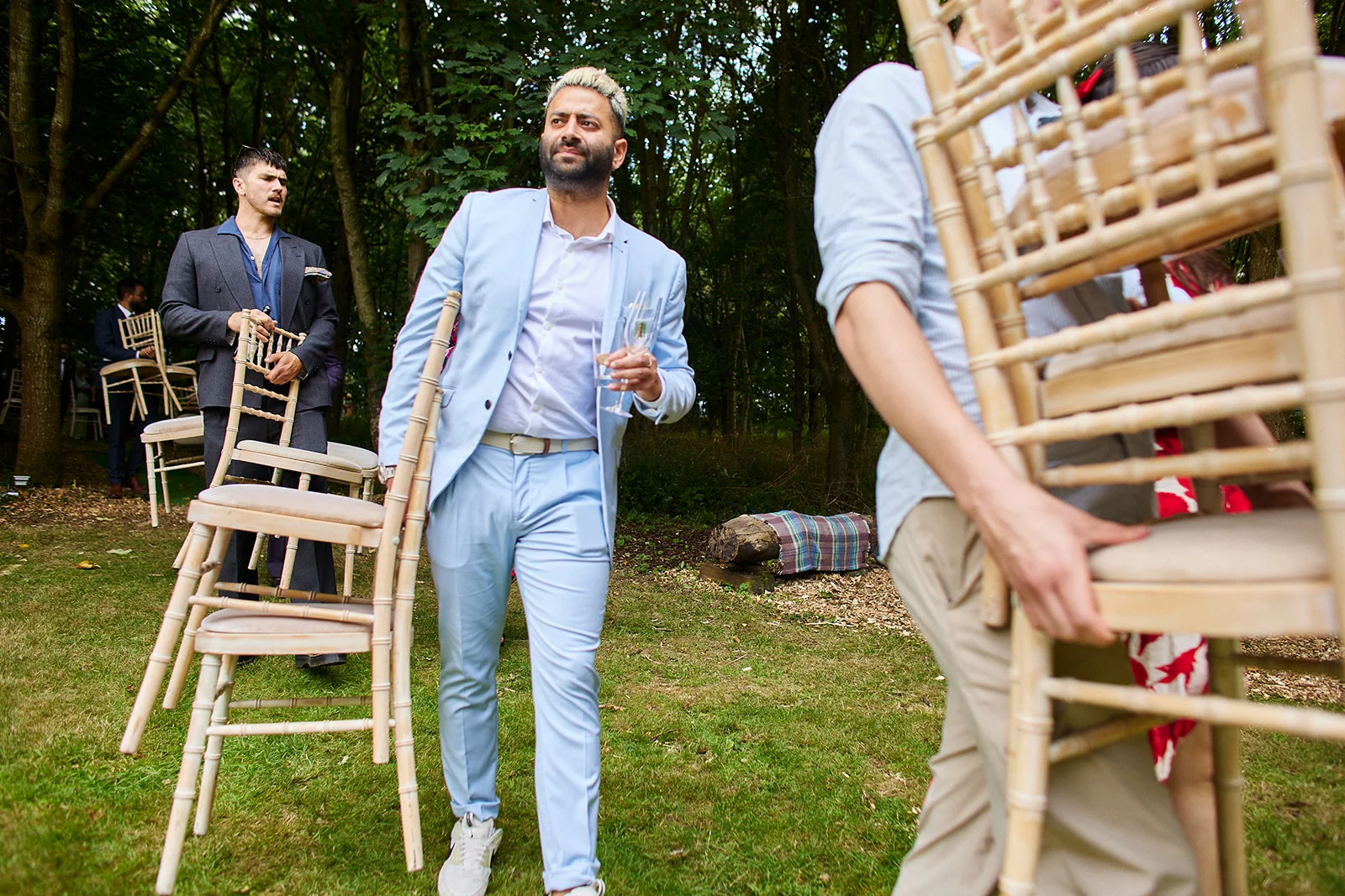 A groom in a light blue suit carrying a stack of chairs through a woodland setting during an outdoor wedding.