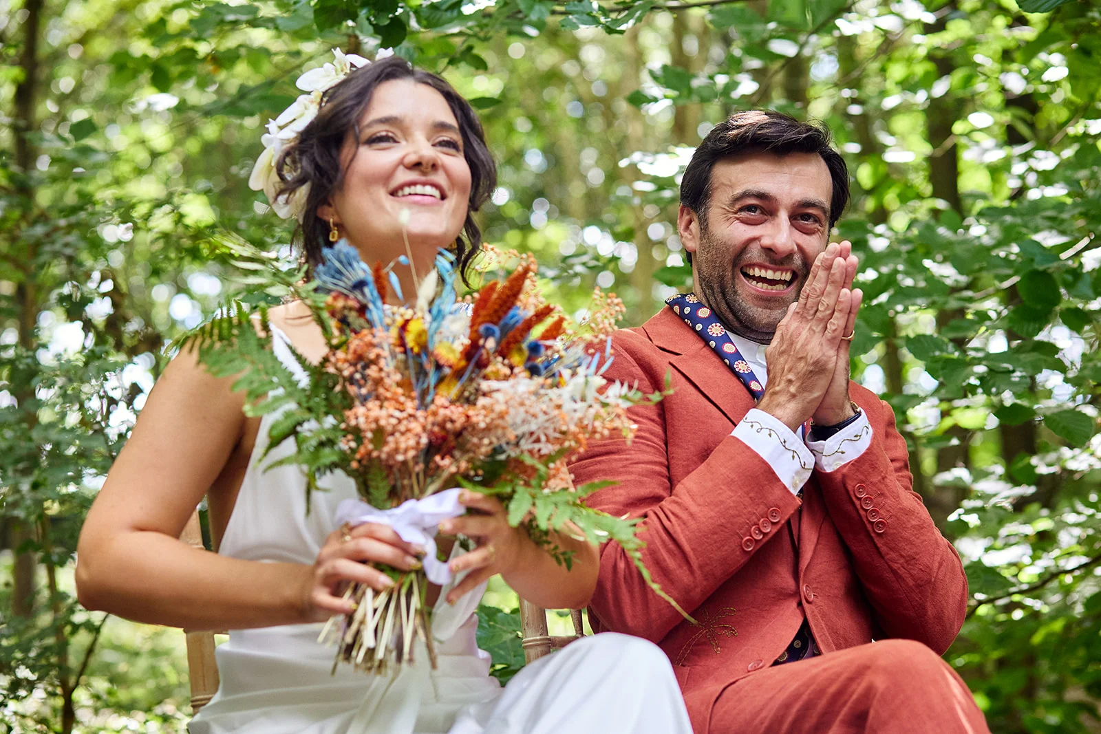 A joyful couple laughs together outdoors in a sunlit woodland setting during their wedding ceremony.