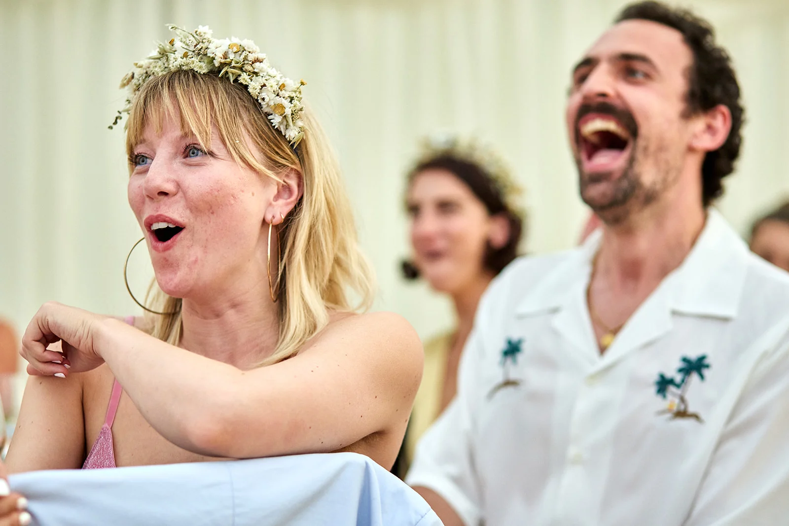 A joyous bride wearing a floral crown and hoop earrings laughs alongside a laughing groom in a patterned open-collared shirt.