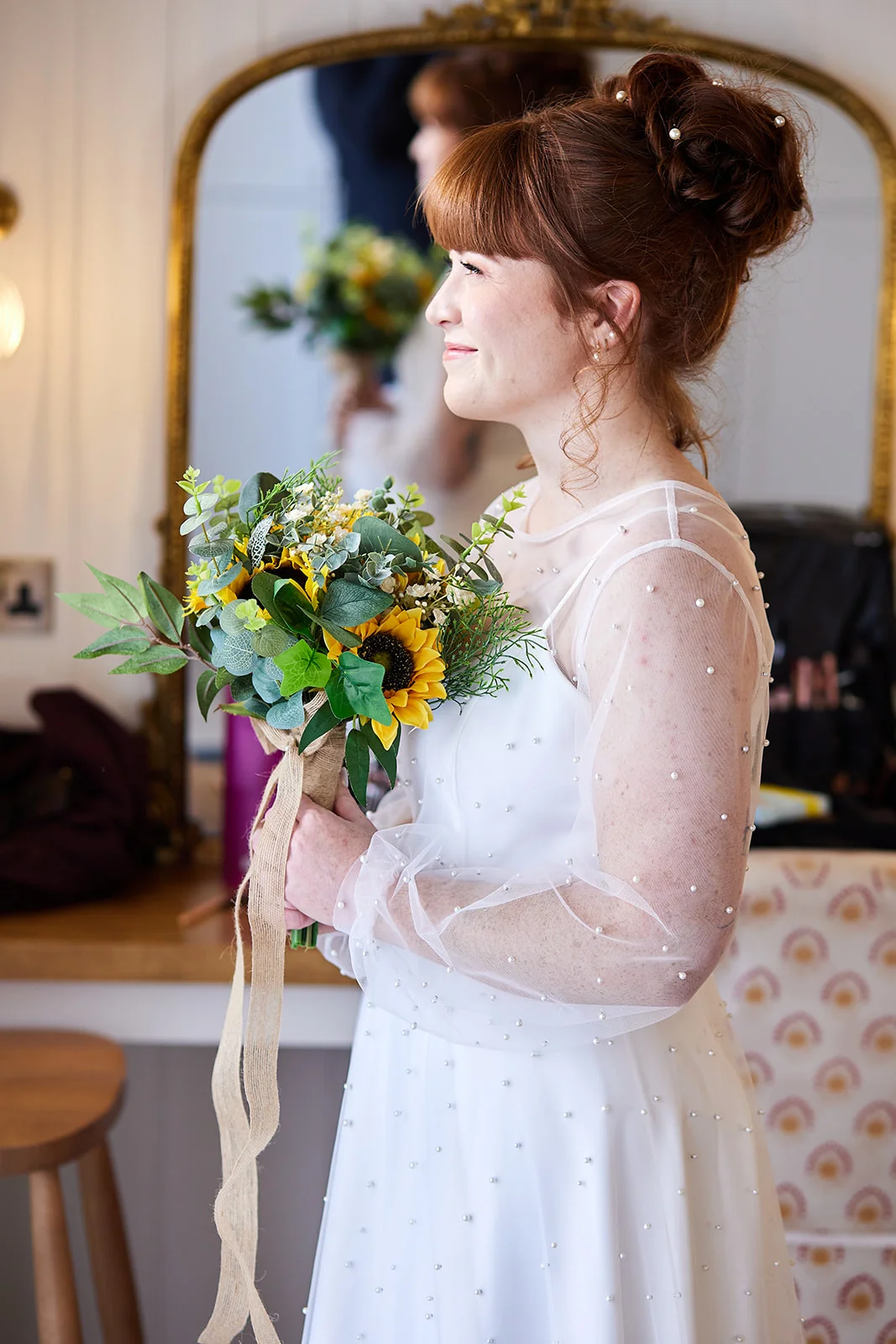 A profile portrait of a smiling bride with red hair tied in a high bun decorated with pearls, wearing a sheer long-sleeved dress covered in pearls while holding a sunflower bouquet.