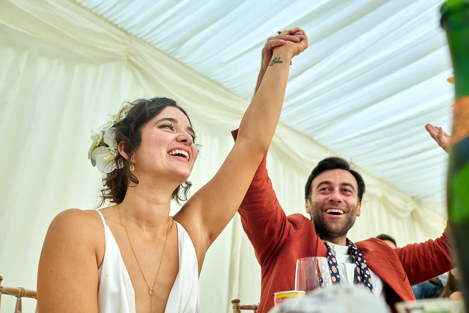 A joyful newlywed couple celebrate together under a draped marquee ceiling, holding their joined hands triumphantly in the air while laughing.