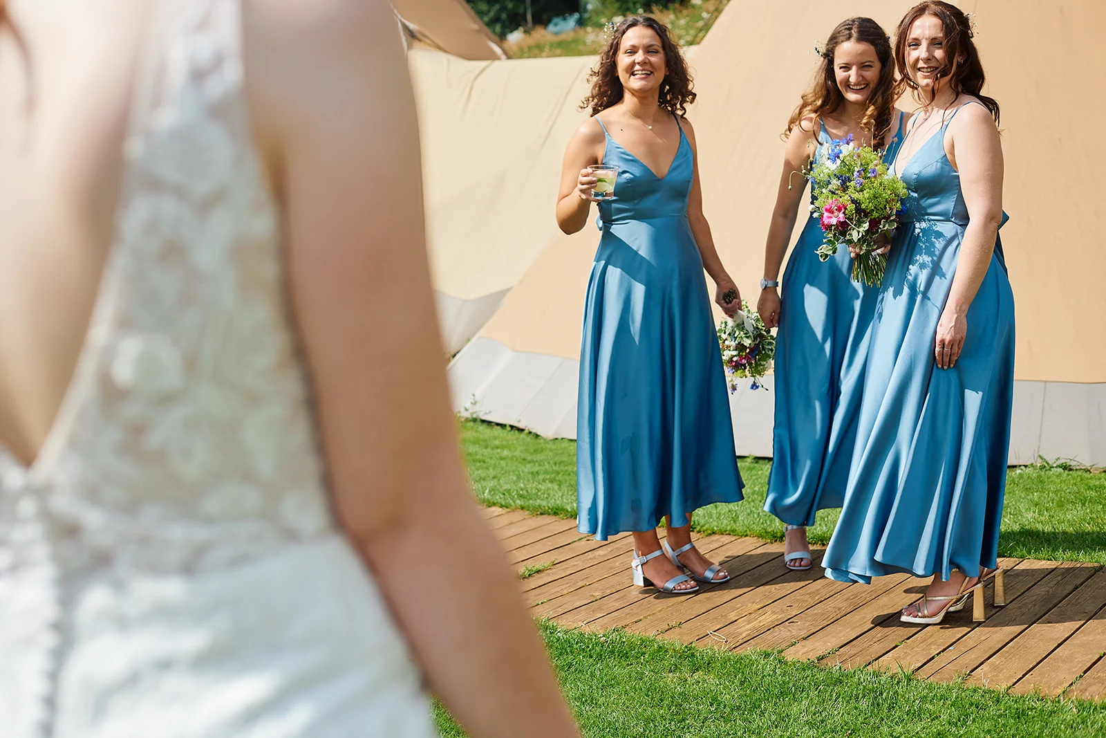 Three smiling bridesmaids in blue satin dresses react with joy during a first look moment outside a marquee wedding venue.