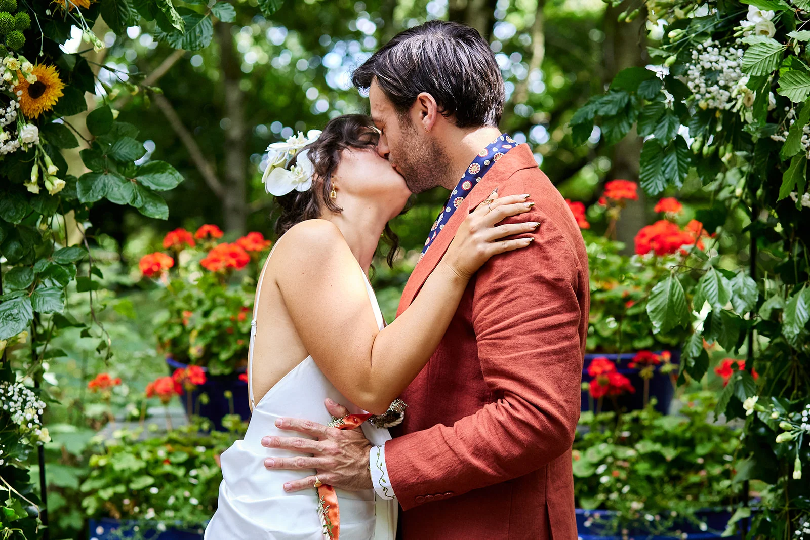 A newlywed couple share a tender kiss framed by a lush floral arch in an outdoor garden setting.