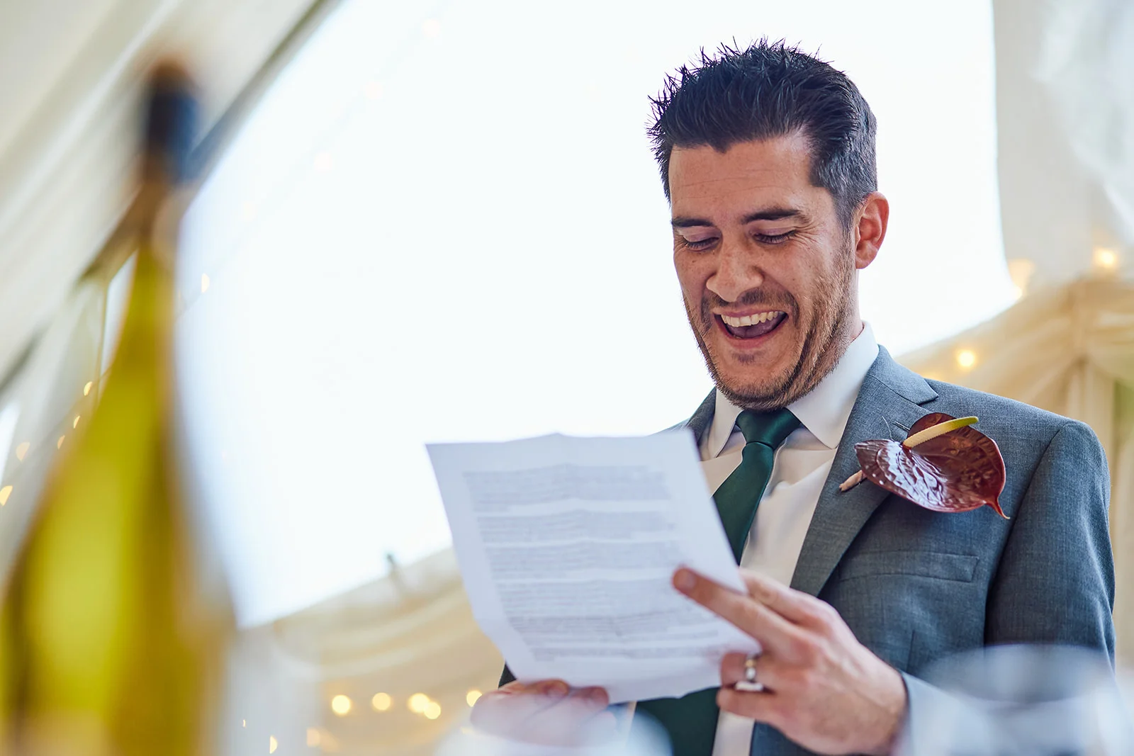 A groom in a grey suit and green tie laughs heartily while reading a speech from a printed paper during the wedding reception.
