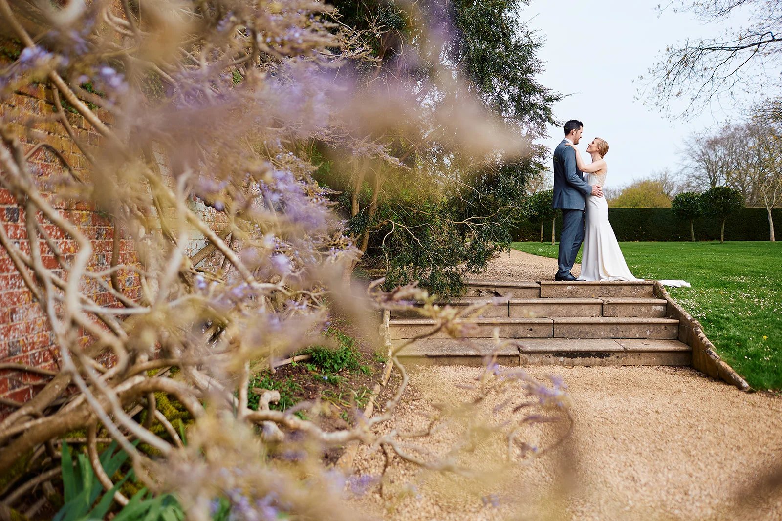 A newlywed couple embraces on stone steps in a manicured garden, framed by flowering wisteria in the foreground.