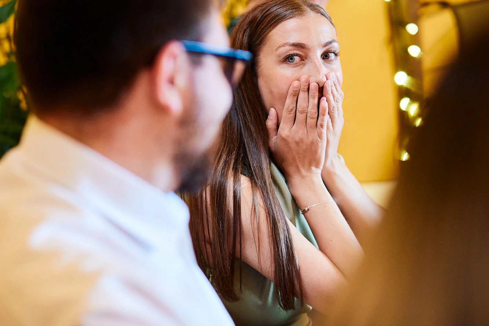 A wedding guest reacts with surprise and laughter during the speeches, holding her hands over her mouth while sitting at a reception table.