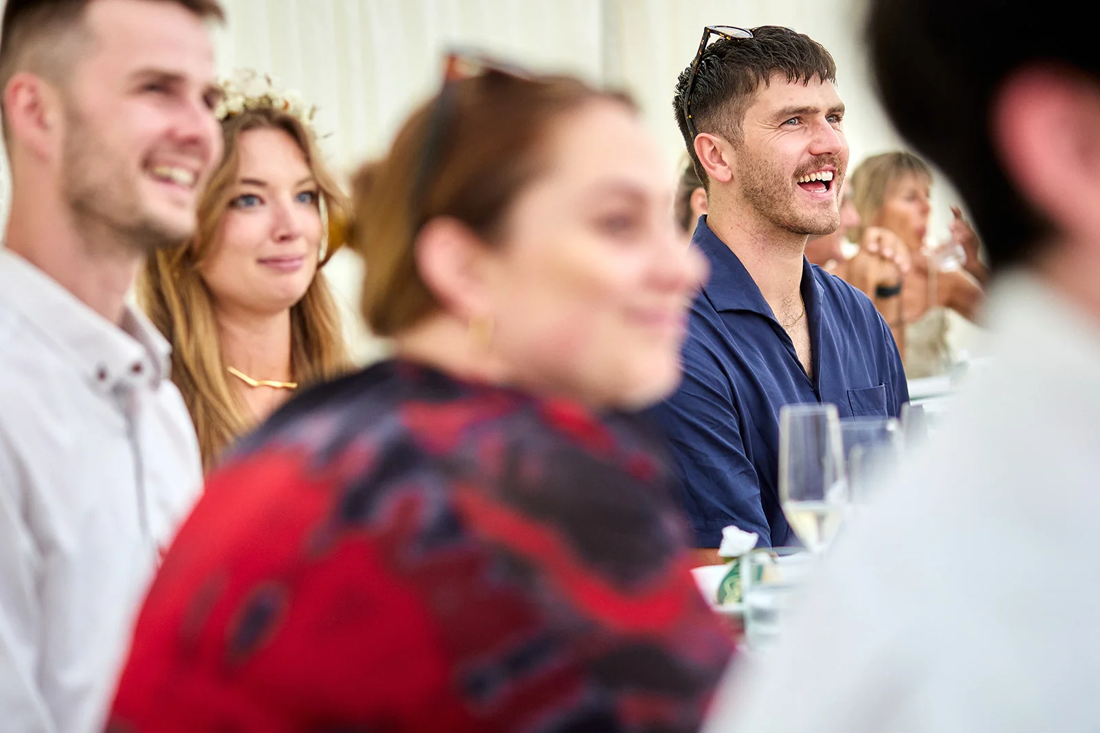 Wedding guests sit and laugh together during the wedding breakfast speeches, captured in a candid shot with natural lighting.