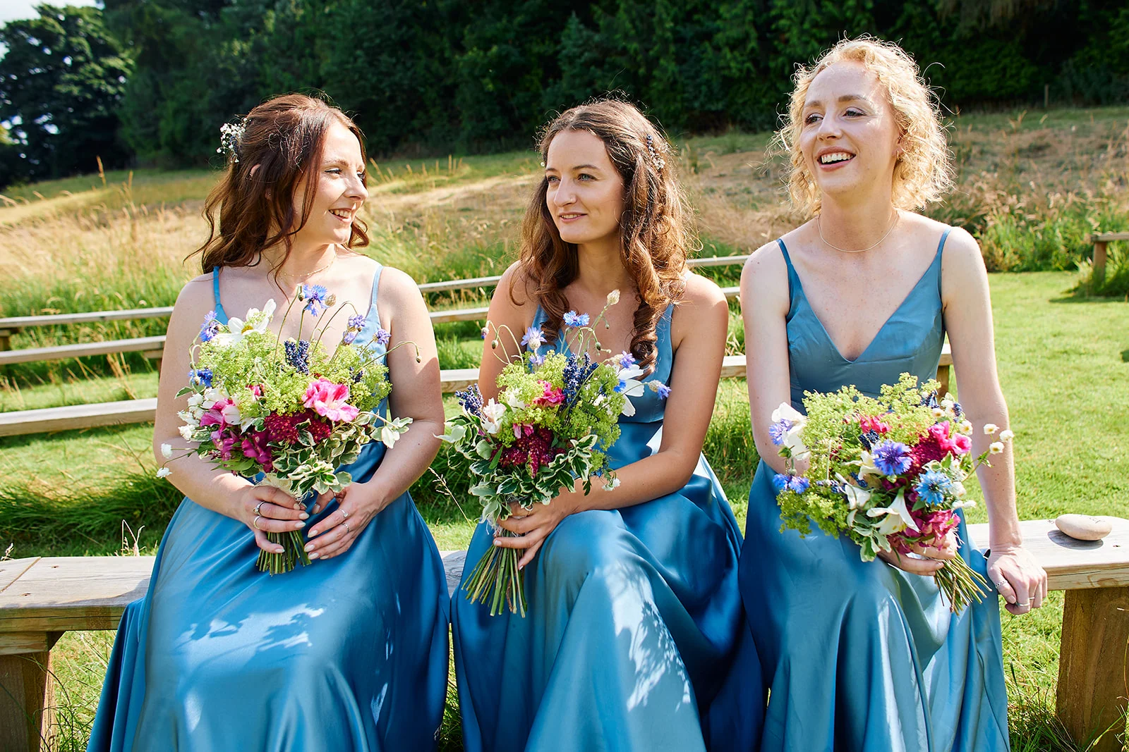 Three smiling bridesmaids in matching satin dusty blue dresses sit outdoors on wooden benches, holding vibrant wild-flower bouquets.