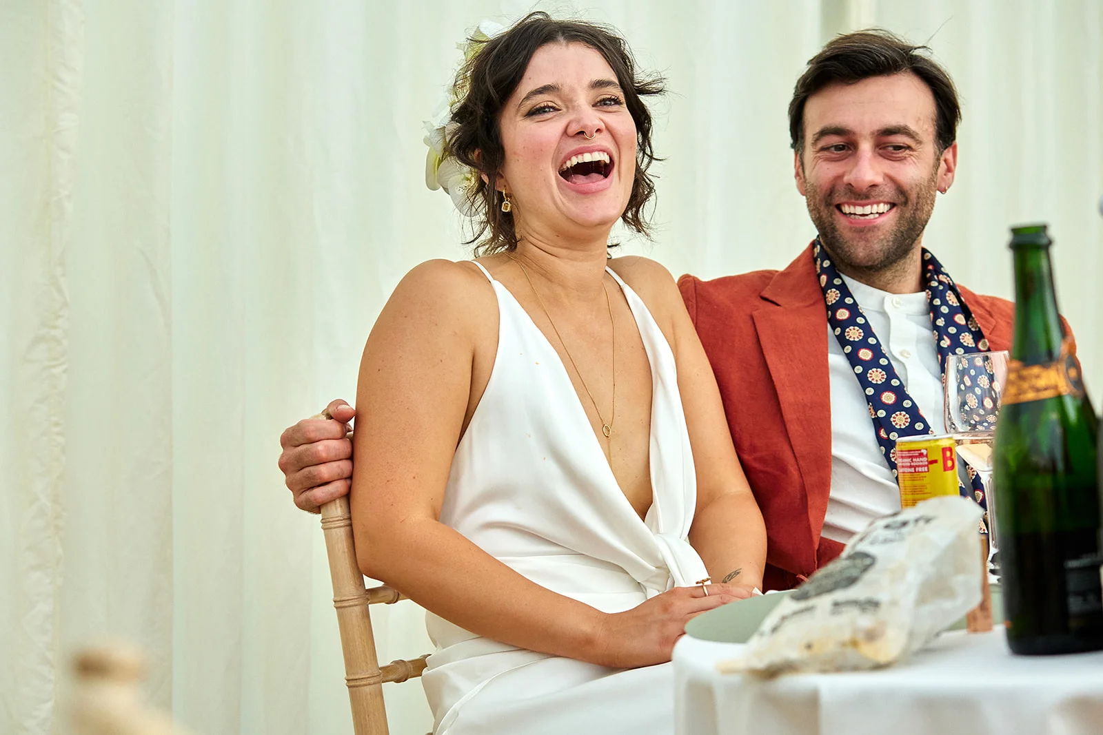 A joyful bride with a white dress and flowers in her hair laughs heartily while sitting next to her groom in a rust-coloured suit during the wedding reception.