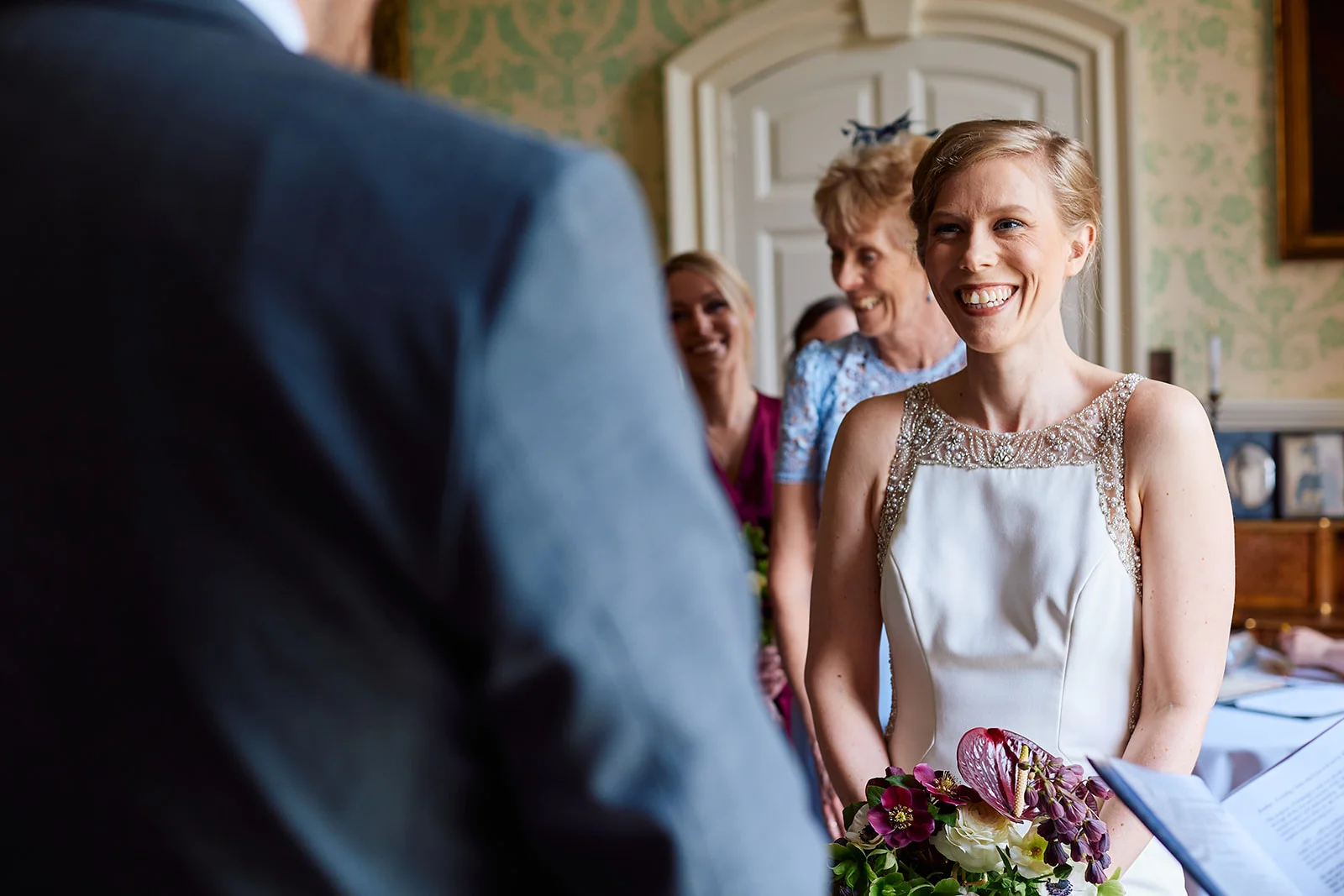 A joyful bride smiles broadly at the groom during their ceremony, holding a vibrant bouquet while guests look on happily in the background.