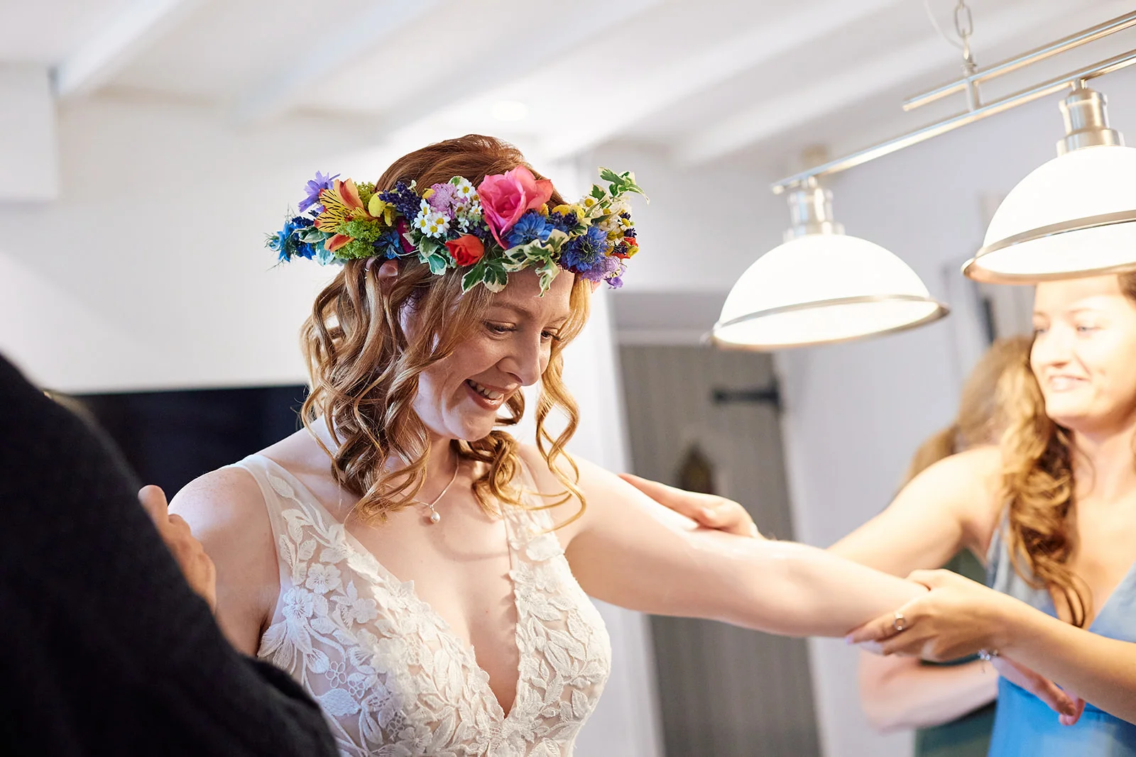 A smiling bride wearing a lace v-neck dress and a vibrant fresh flower crown gets her arm cared for by bridesmaids during morning preparations.