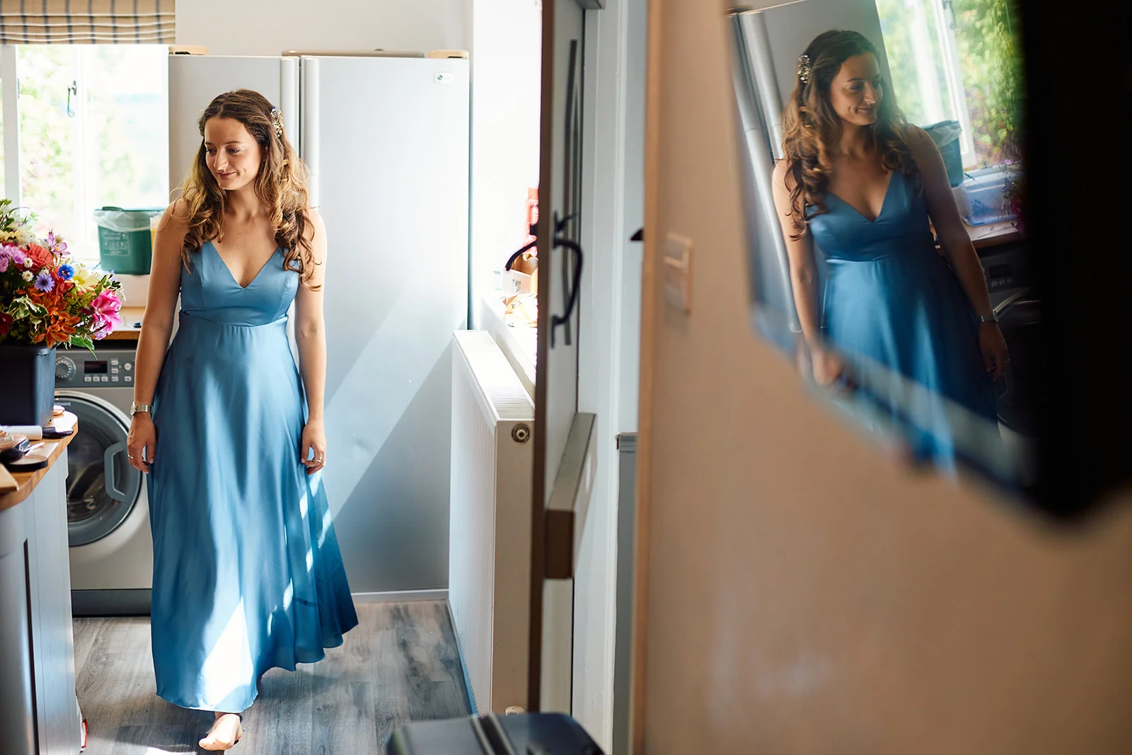 A bridesmaid in a blue satin dress is captured in a candid mirror reflection as she gets ready indoors.