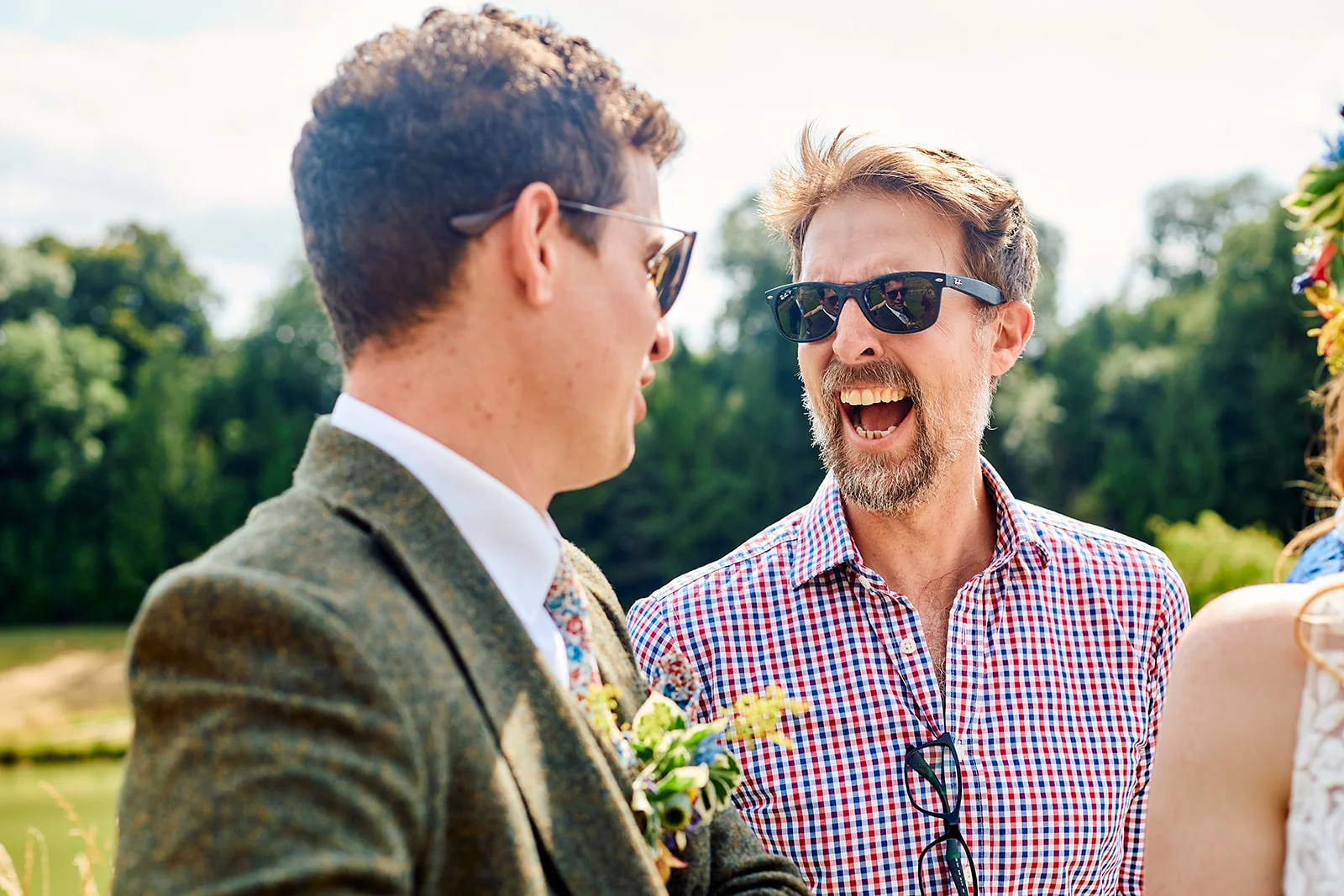 Two male wedding guests wearing sunglasses and casual or tweed tailoring share a joyful, laughing conversation outdoors in bright sunlight.