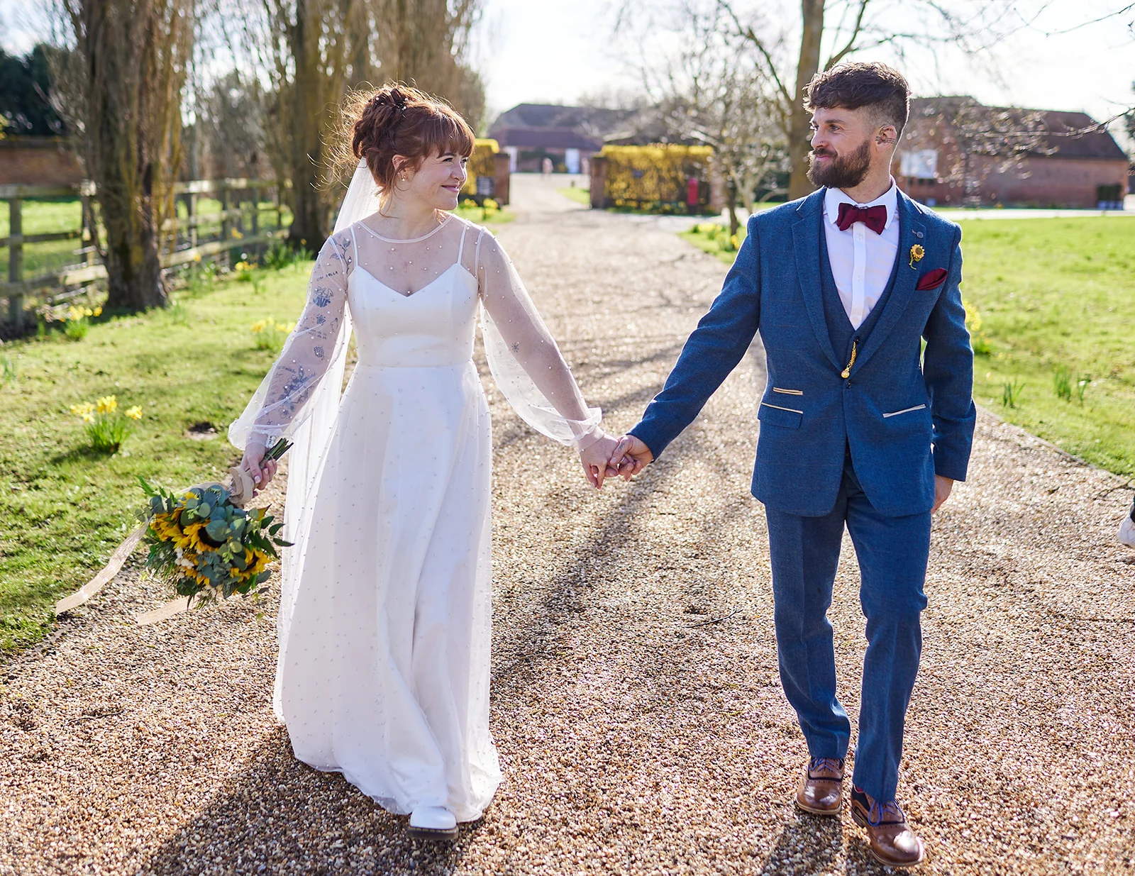 A bride and groom walk hand in hand down a gravel driveway lined with trees, sharing a loving glance in the bright spring sunshine.