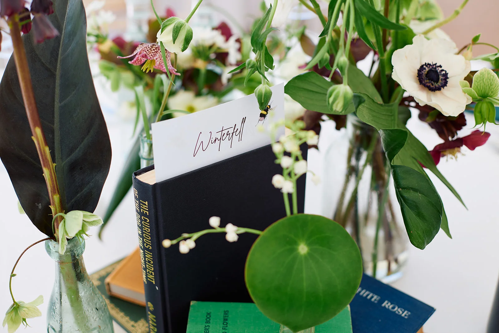 A close-up of a literary-themed wedding table name card featuring the word Winterfell nestled among stacked vintage books and delicate bud vases filled with white anemones and greenery.