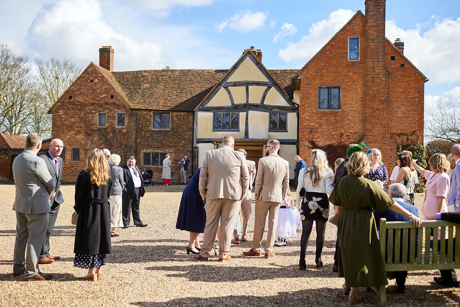 Wedding guests mingle in the gravel courtyard of a historic red brick manor house on a sunny afternoon.