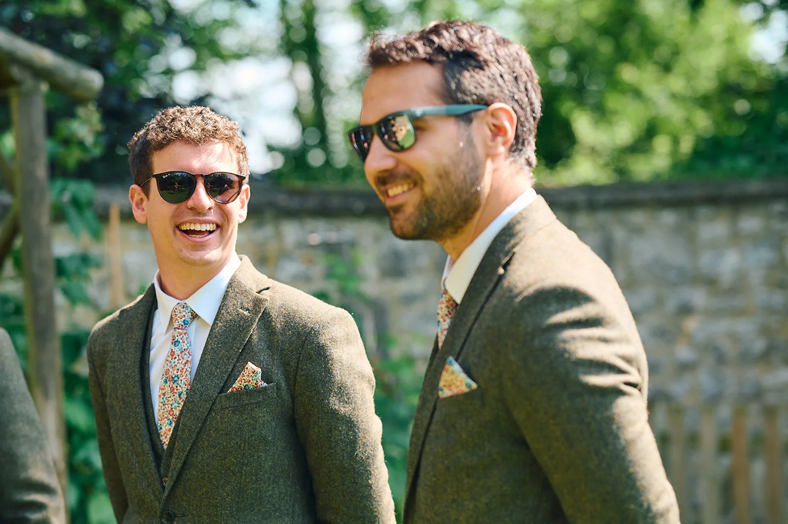 Two smiling groomsmen wearing matching tweed suits and floral ties stand outdoors in bright sunlight.