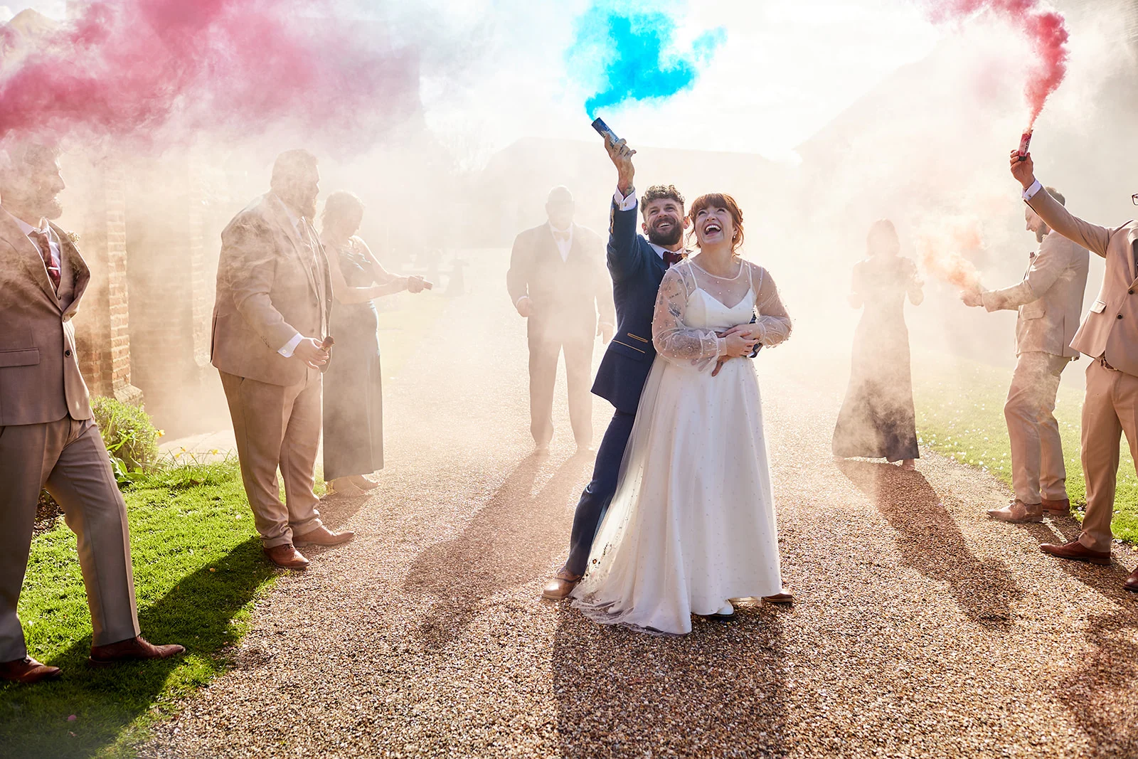 A newlywed couple laughs joyfully as they are surrounded by vibrant pink and blue smoke bombs held by their wedding party outdoors.