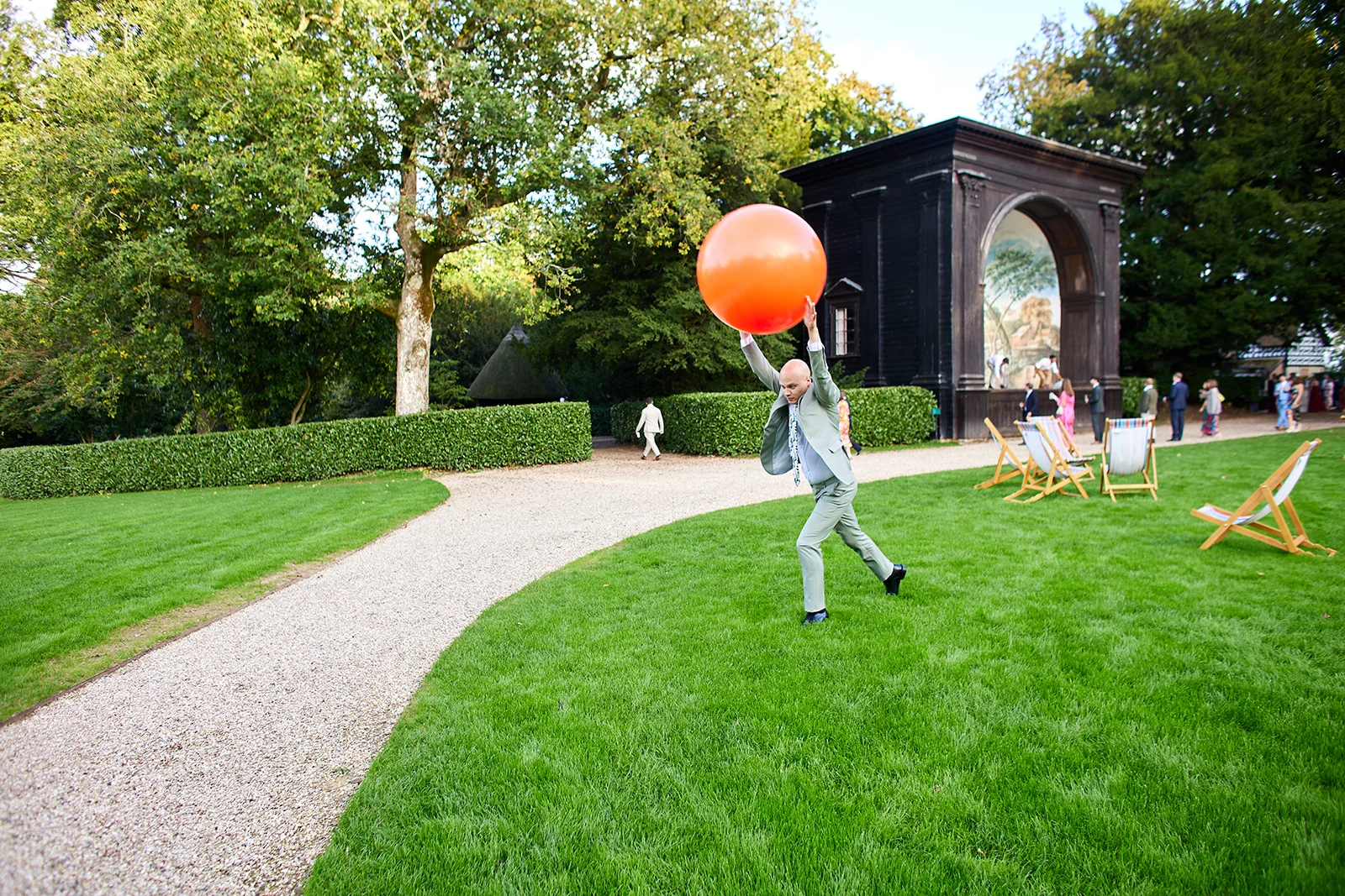 A guest in a green suit playfully holds a large orange balloon on the manicured lawn of a country house wedding venue during a sunny afternoon.