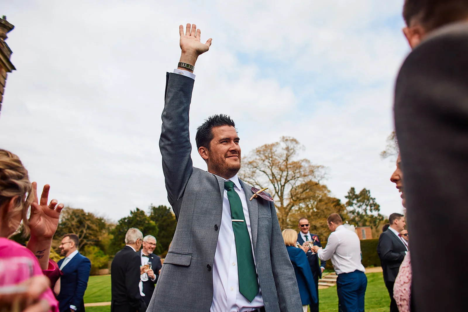 A groom in a grey suit and green tie raises his hand to wave during an outdoor wedding drinks reception in a manicured garden.