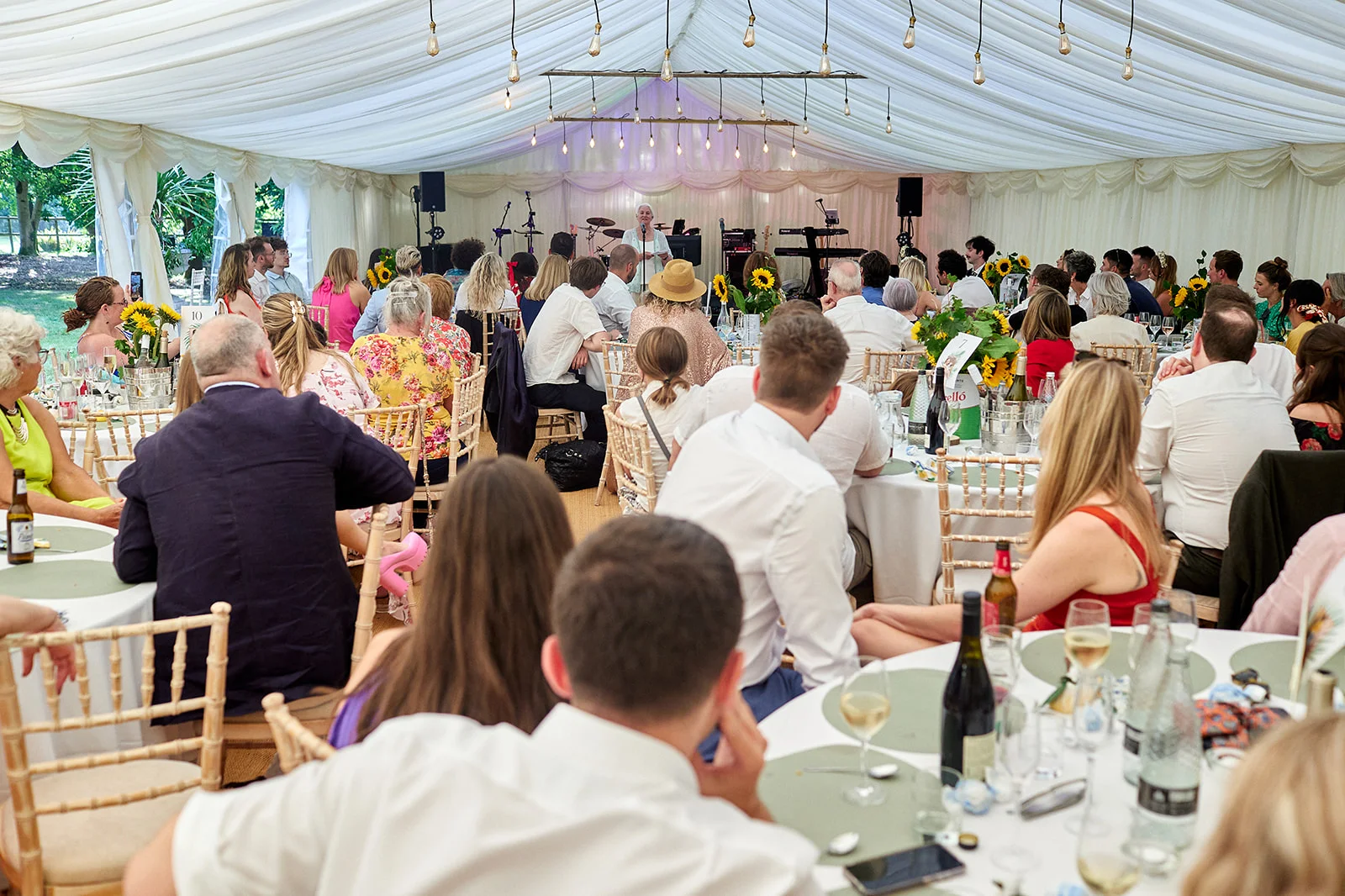 A wide shot shows wedding guests seated at round tables inside a white draped marquee, listening to a singer performing at the front.