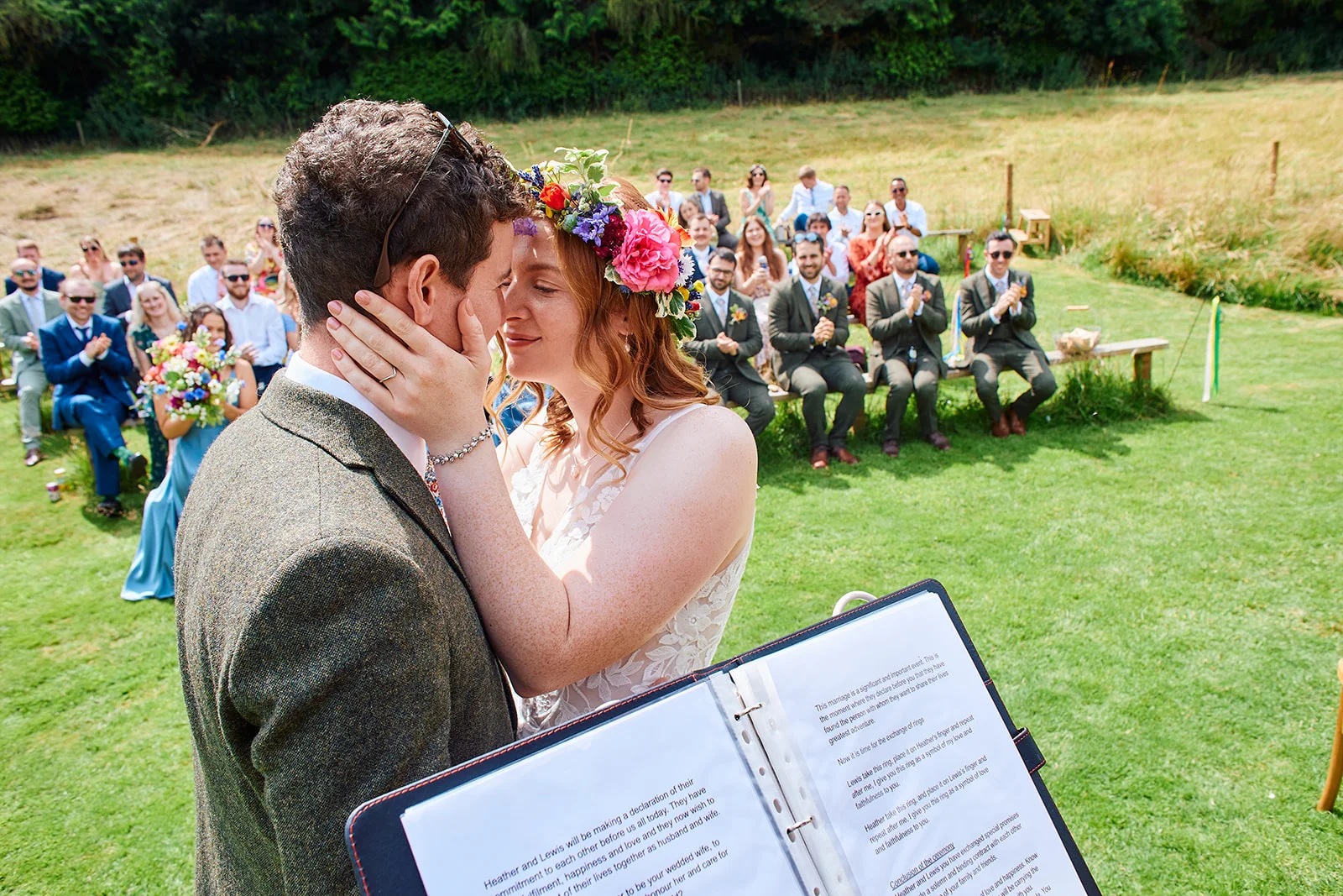 A bride wearing a colourful floral crown and groom in a tweed suit share an intimate forehead kiss during an outdoor celebrant-led ceremony, with seated guests watching on in a sunny field.