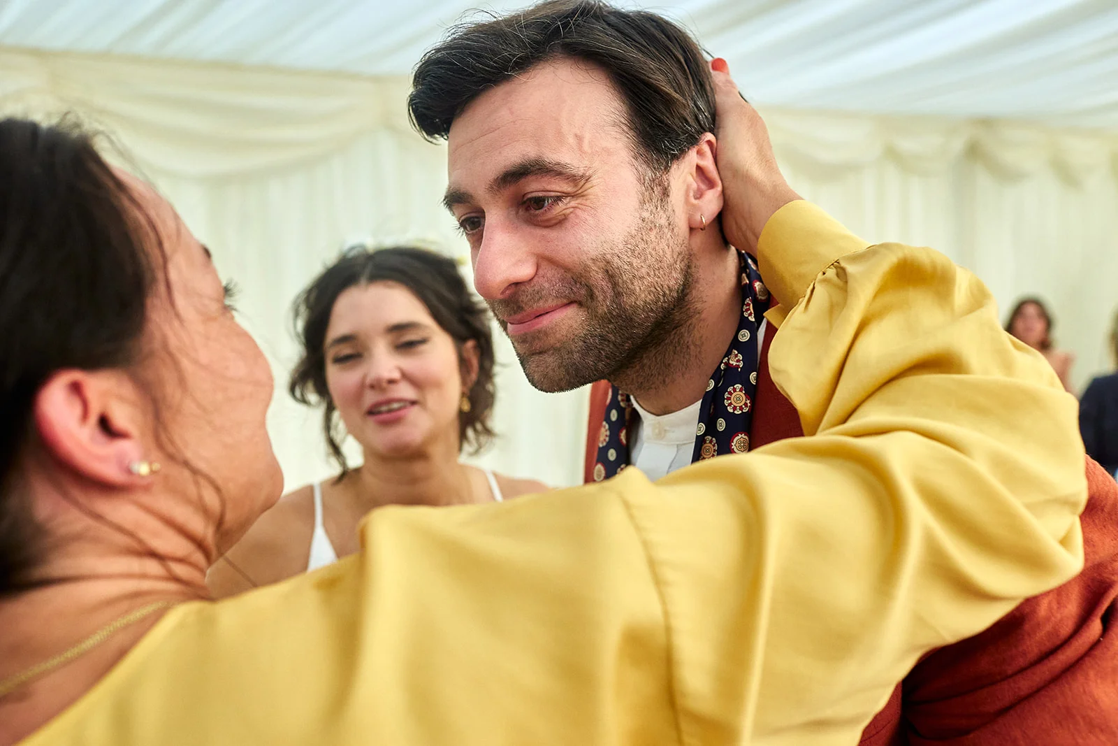 A groom shares a tender, emotional embrace with a guest wearing a yellow blouse inside a marquee during the wedding reception.